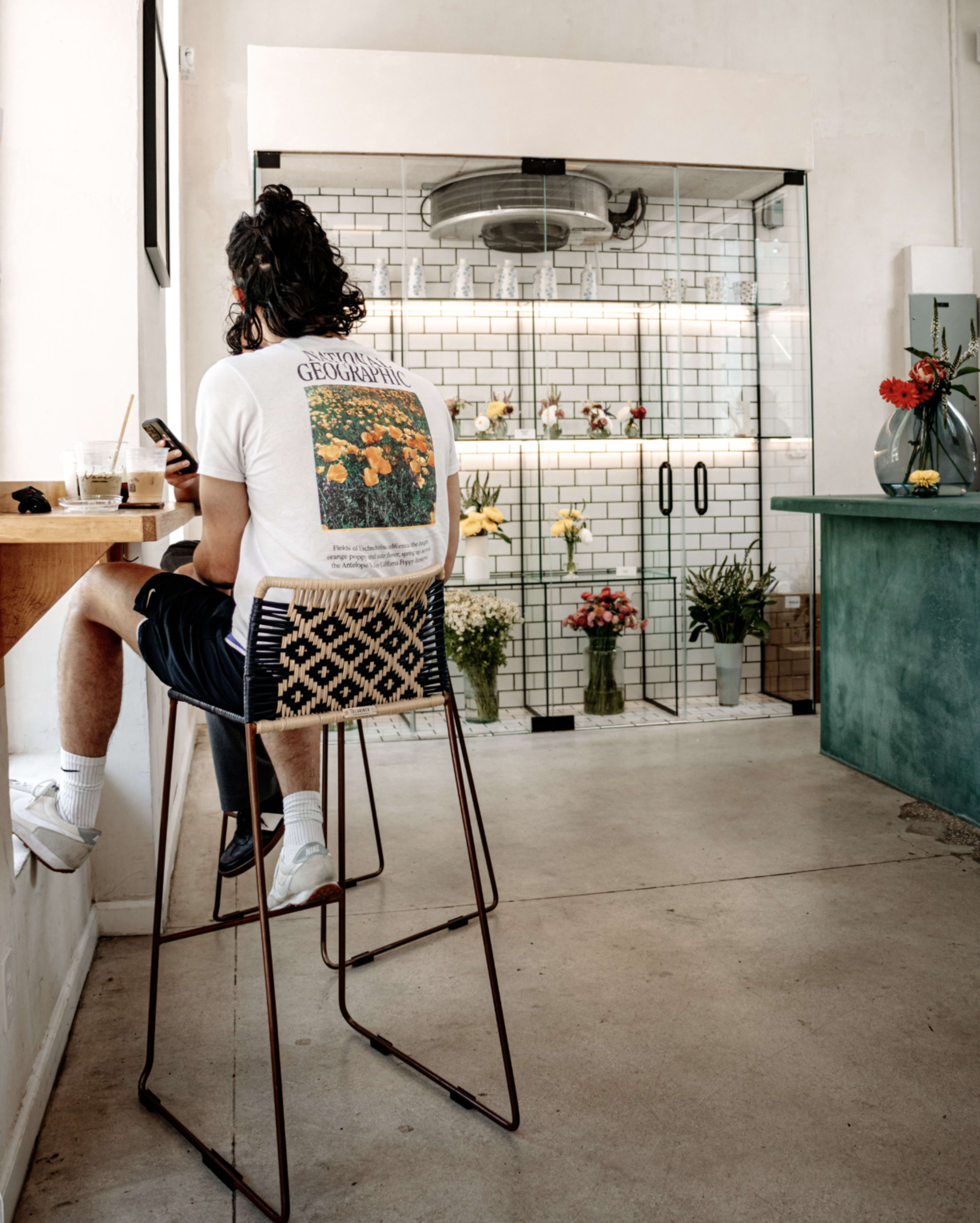 A person sits on a high stool at a counter in a café, looking at their phone while surrounded by potted plants and flowers.