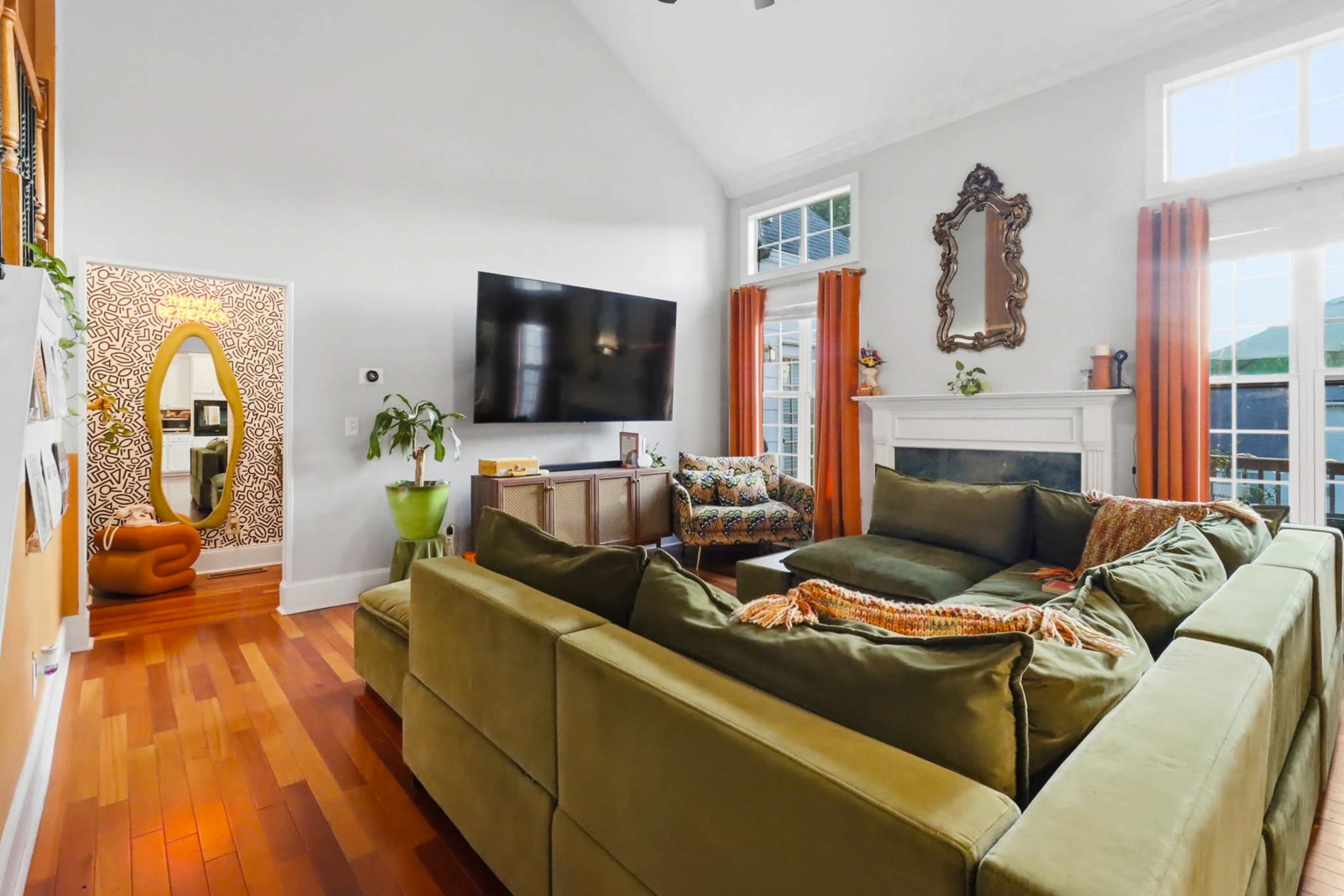 The living room features a large sectional sofa arranged around a flat-screen TV, with sunlight streaming in through tall windows and a decorative mirror in the background.
