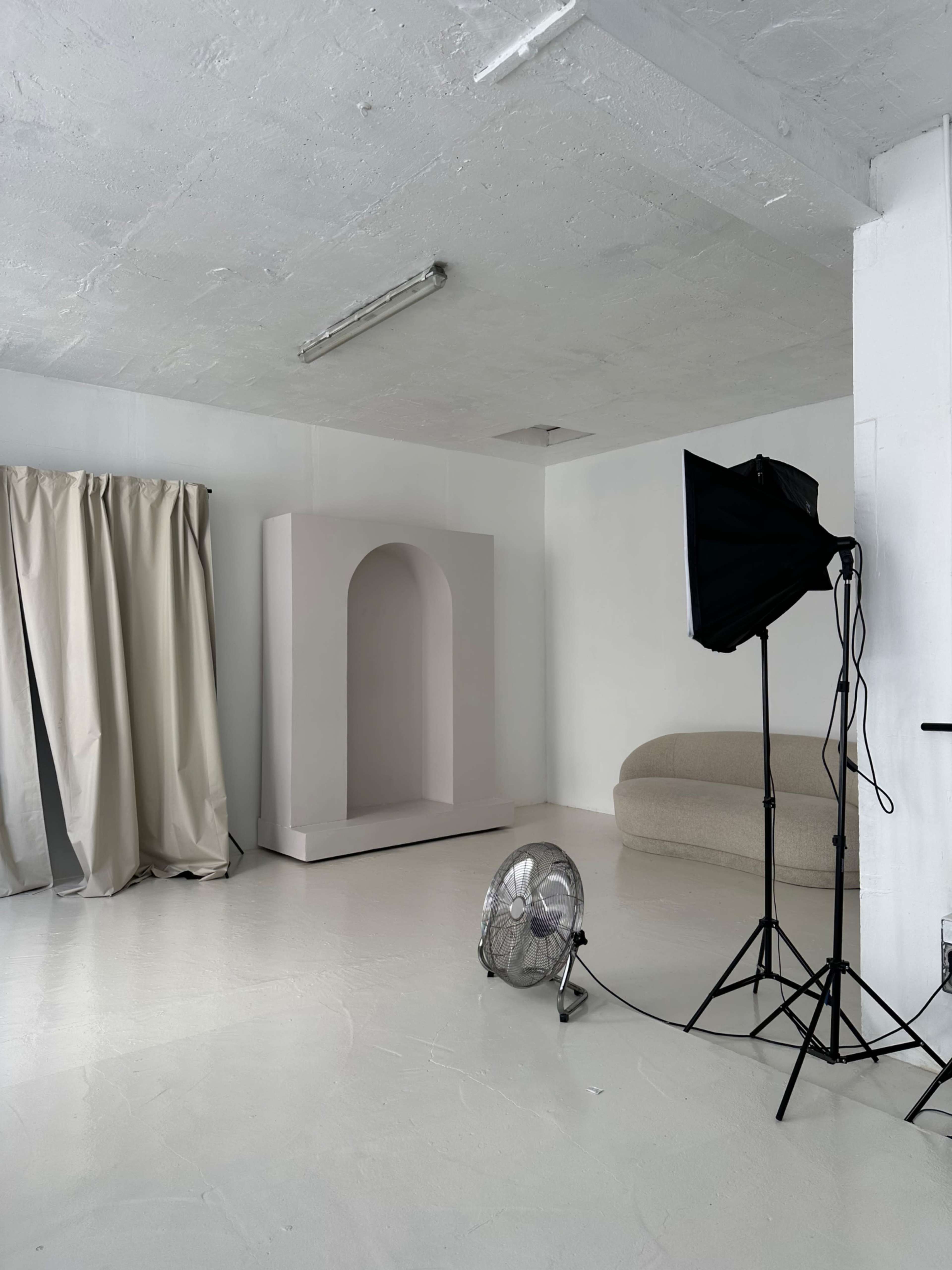 A minimalist studio featuring a large archway structure, a light-colored couch, a fan, and a light stand with softbox, all against a white backdrop.