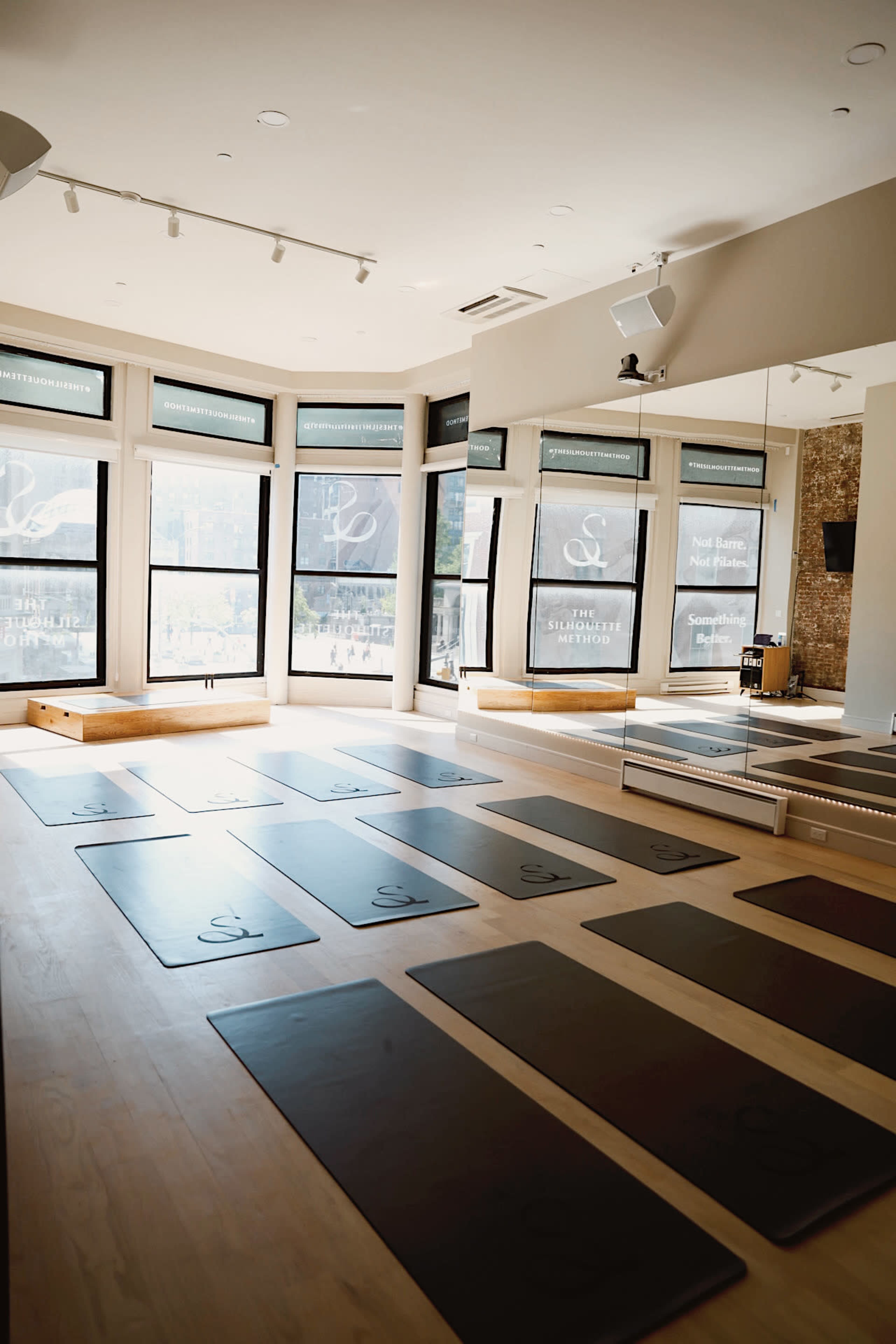 The image shows a well-lit yoga studio with several yoga mats arranged on a wooden floor and large windows featuring promotional signage.