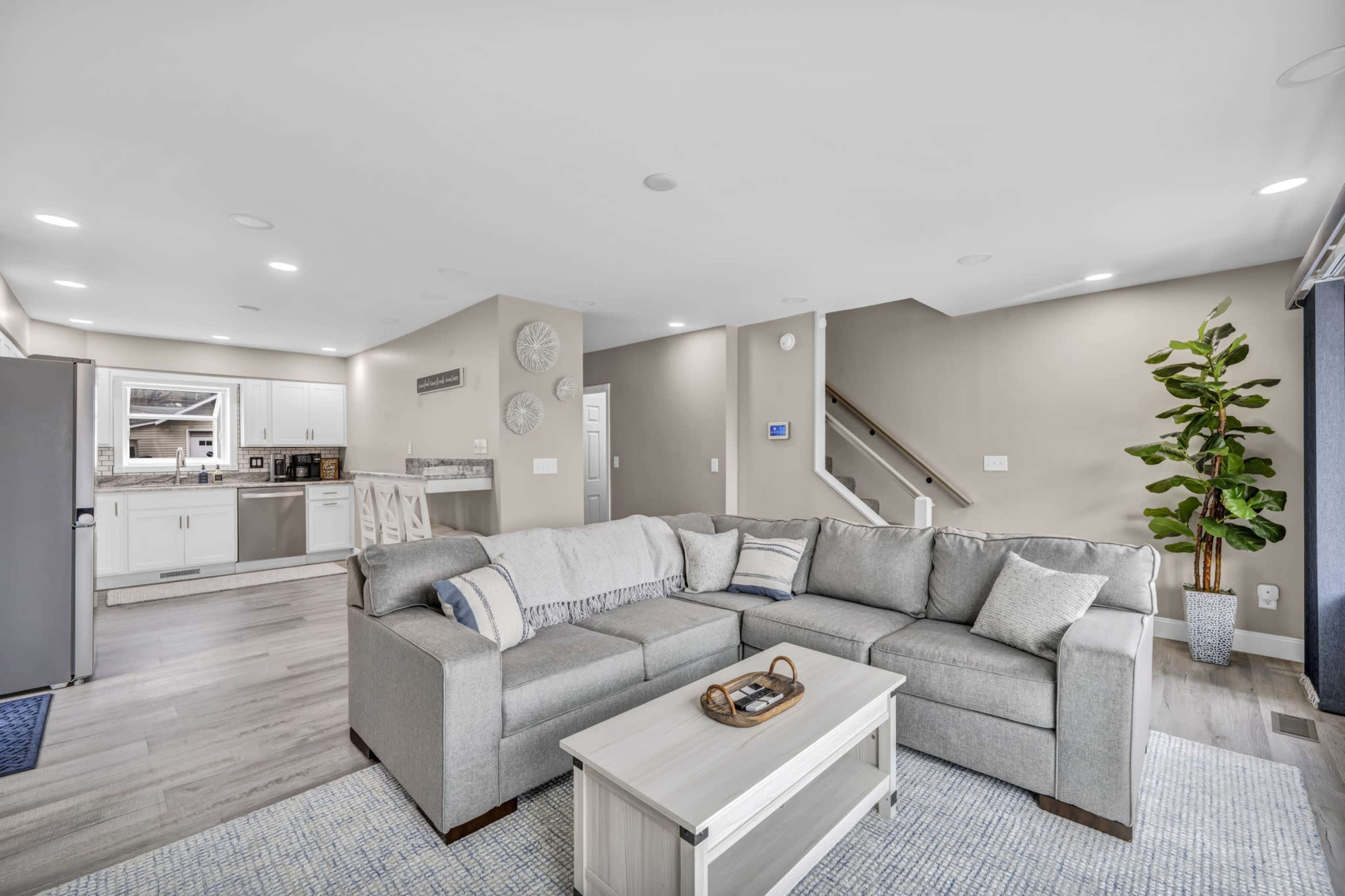 The image shows a modern living room with a gray sectional sofa, a white coffee table, and an open kitchen area in the background.