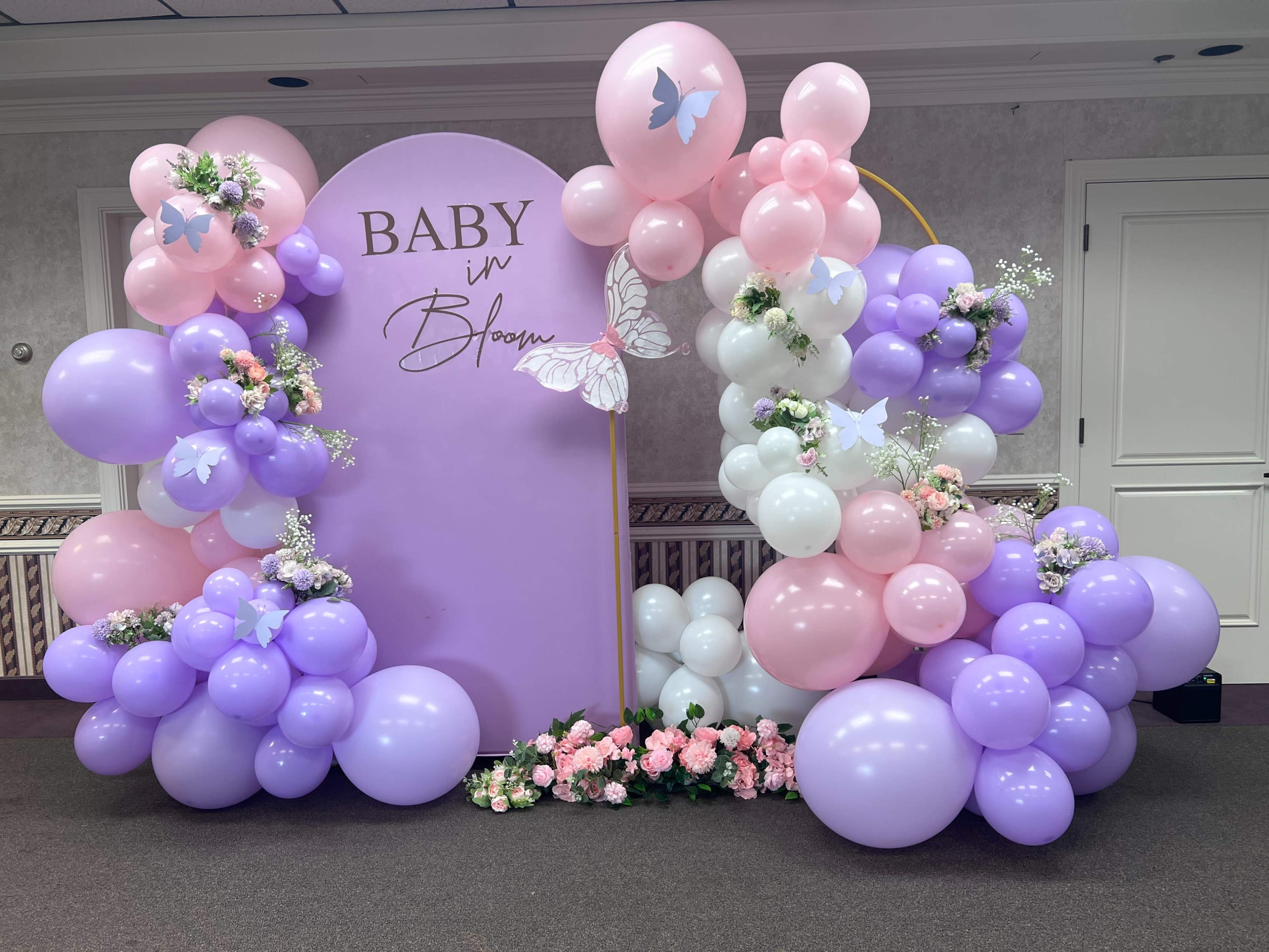 A decorative setup for a baby shower, with an arch made of purple, pink, and white balloons, surrounded by flowers and a central sign that reads "Baby in Bloom."