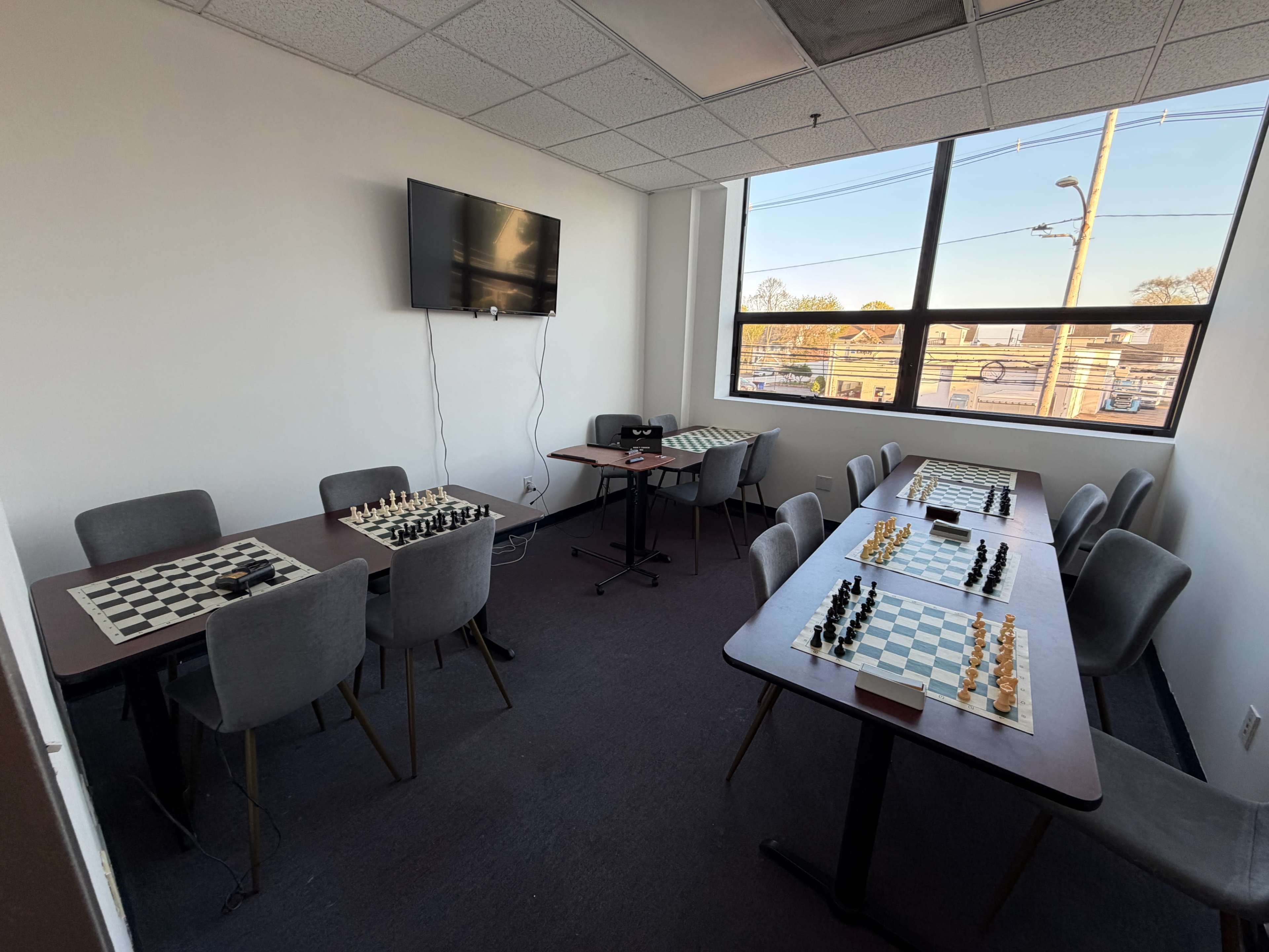 The room contains two tables set up with chess boards and pieces, with a television mounted on the wall and large windows letting in natural light.