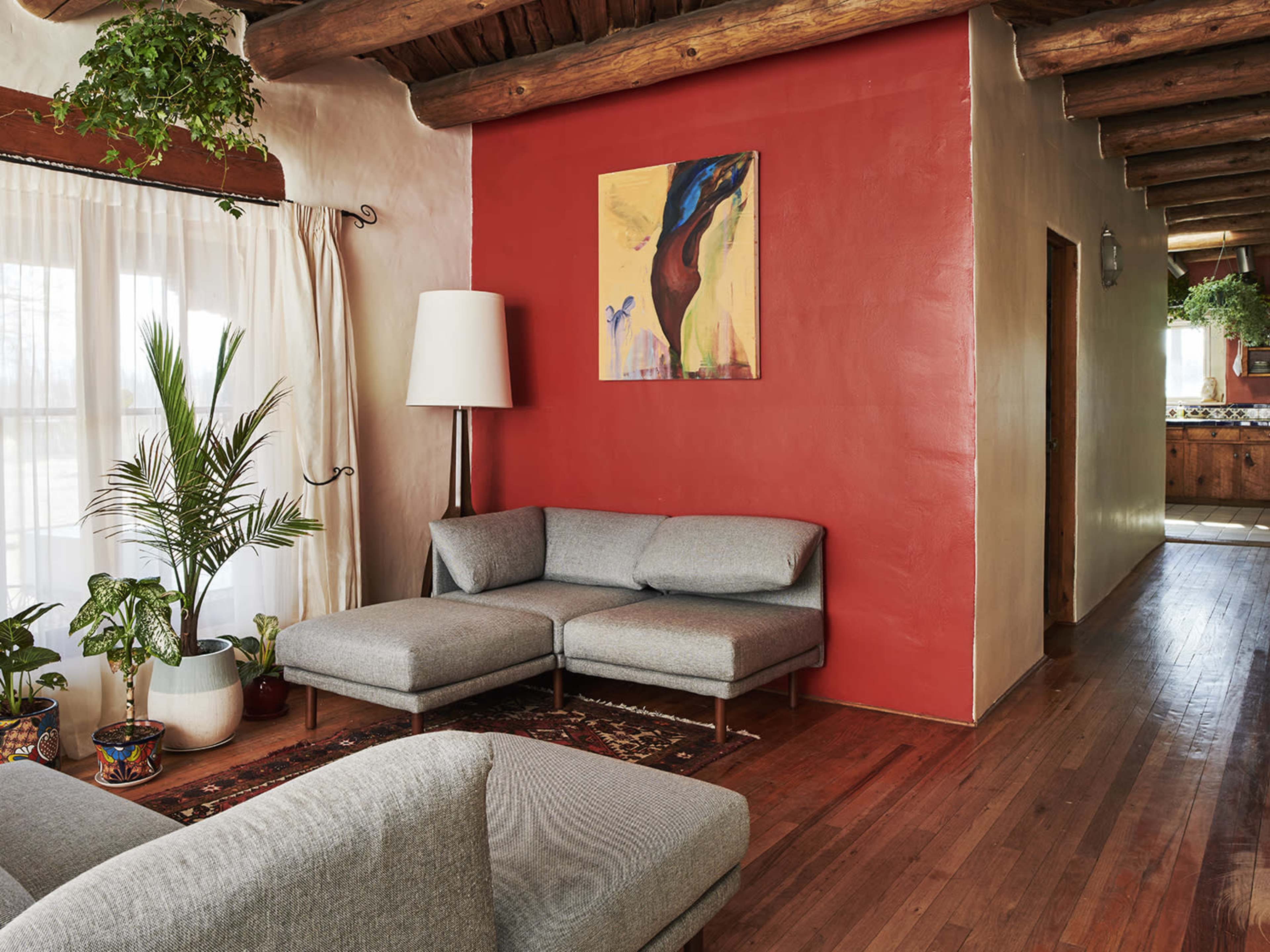A living room with a gray sectional sofa, a tall white lamp, potted plants, and a red accent wall featuring a piece of artwork.