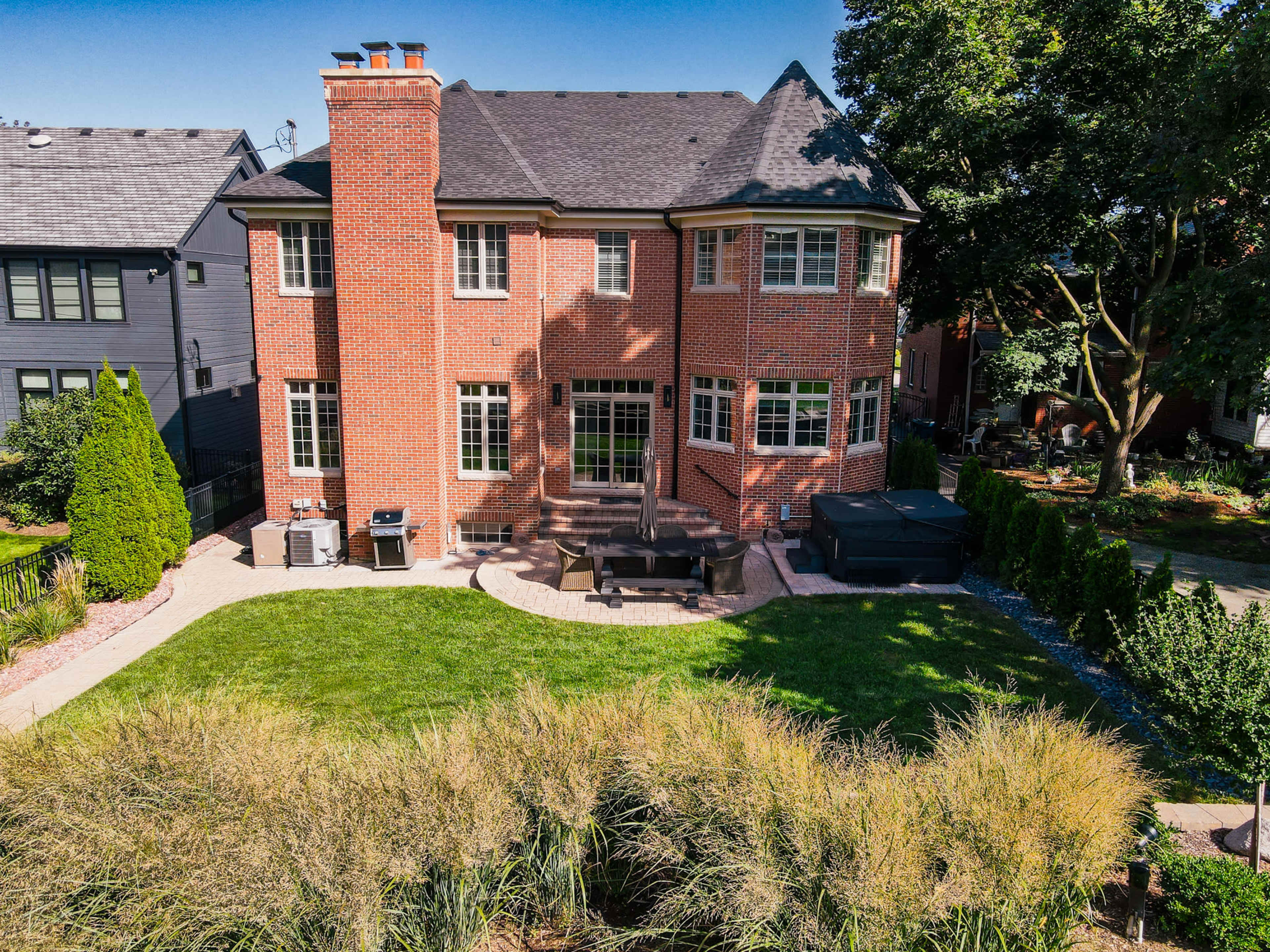 The image shows a two-story brick house with a patio, outdoor furniture, and a hot tub surrounded by landscaped greenery.