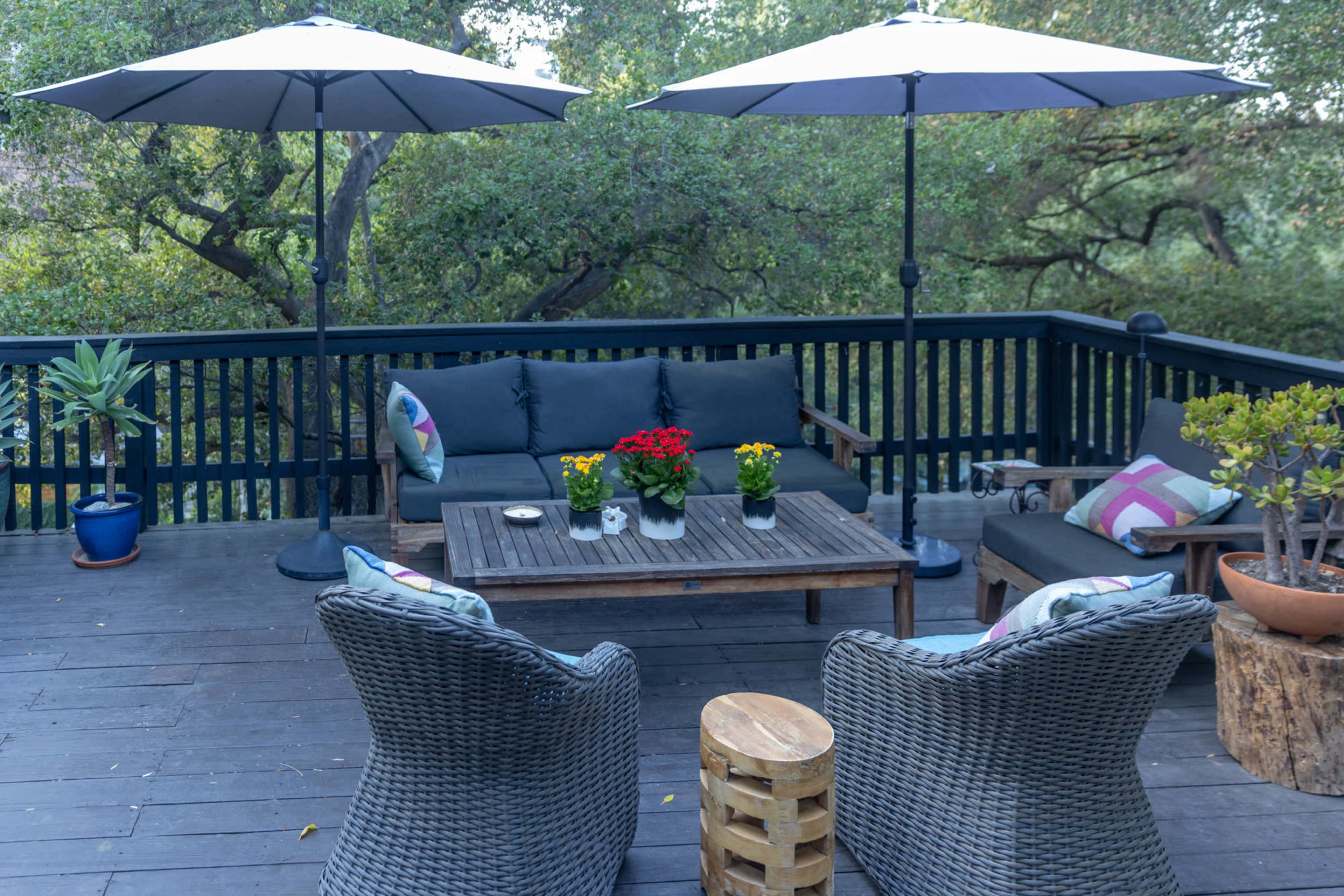The image shows a wooden patio area with two umbrellas, a seating arrangement with cushions, and potted plants on the table and surrounding areas.