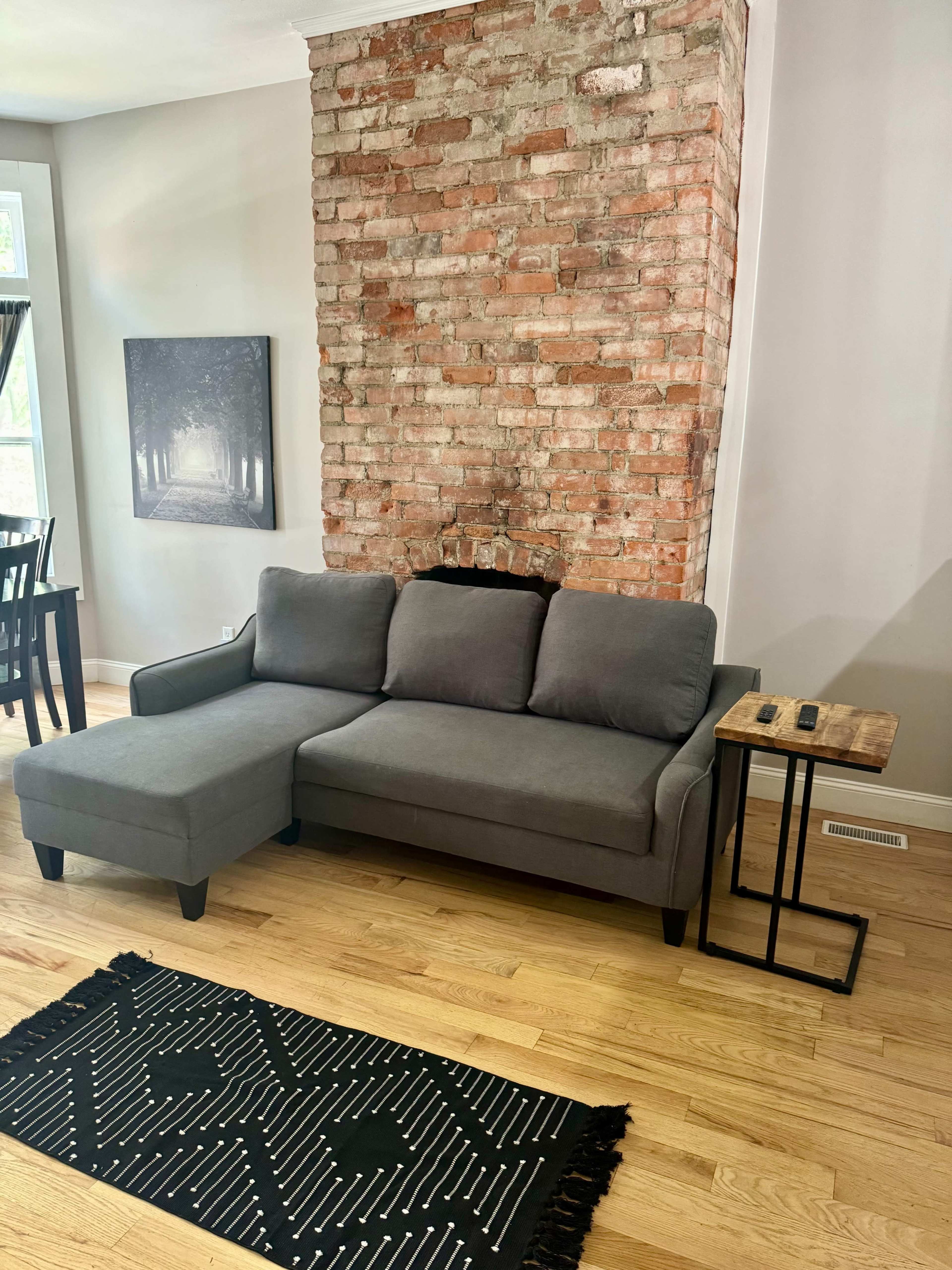 A gray sectional sofa is positioned against a exposed brick wall, accompanied by a small wooden side table and a patterned rug on a hardwood floor.