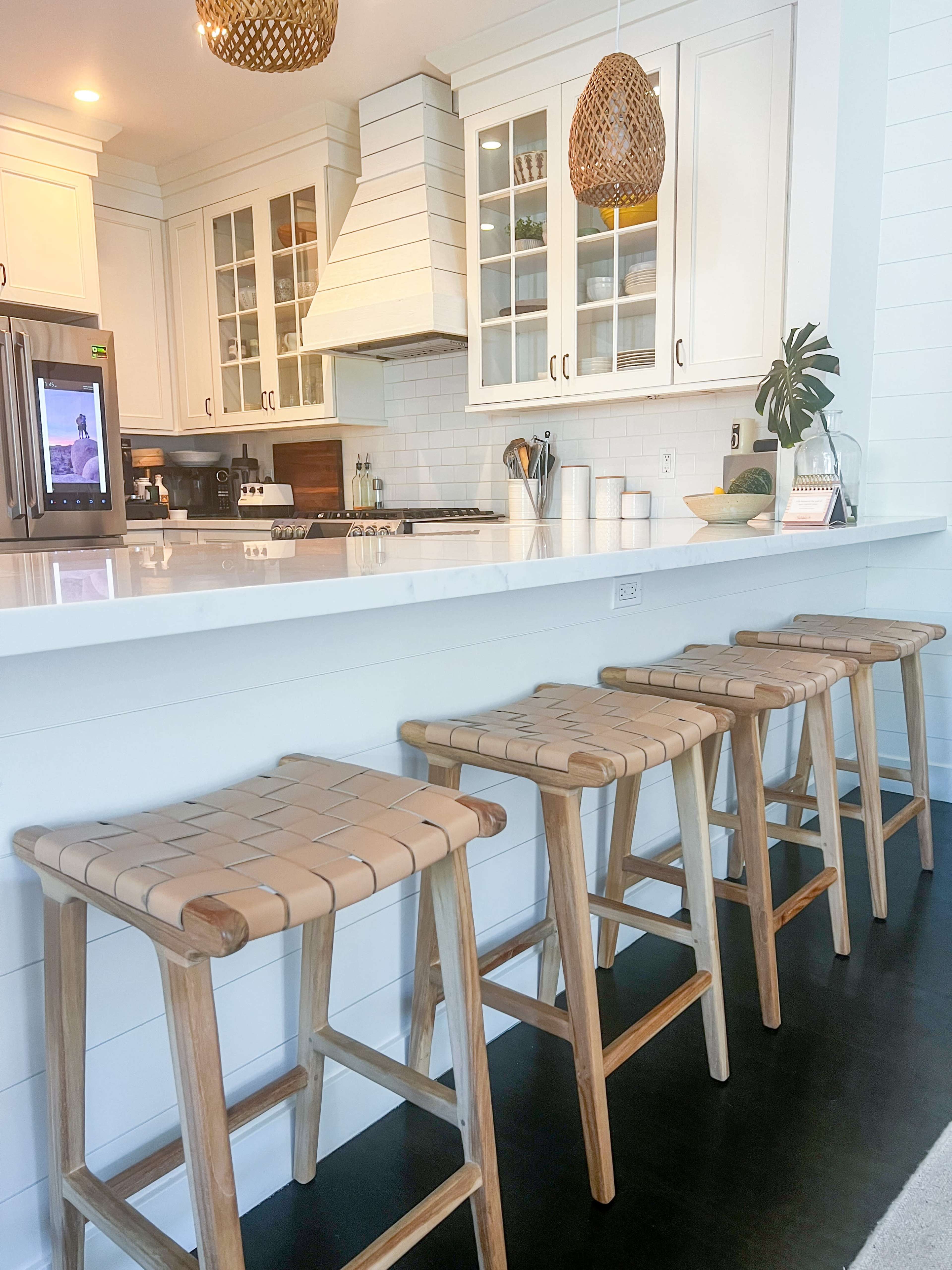 A modern kitchen features a white countertop with four wooden bar stools and hanging pendant lights above.