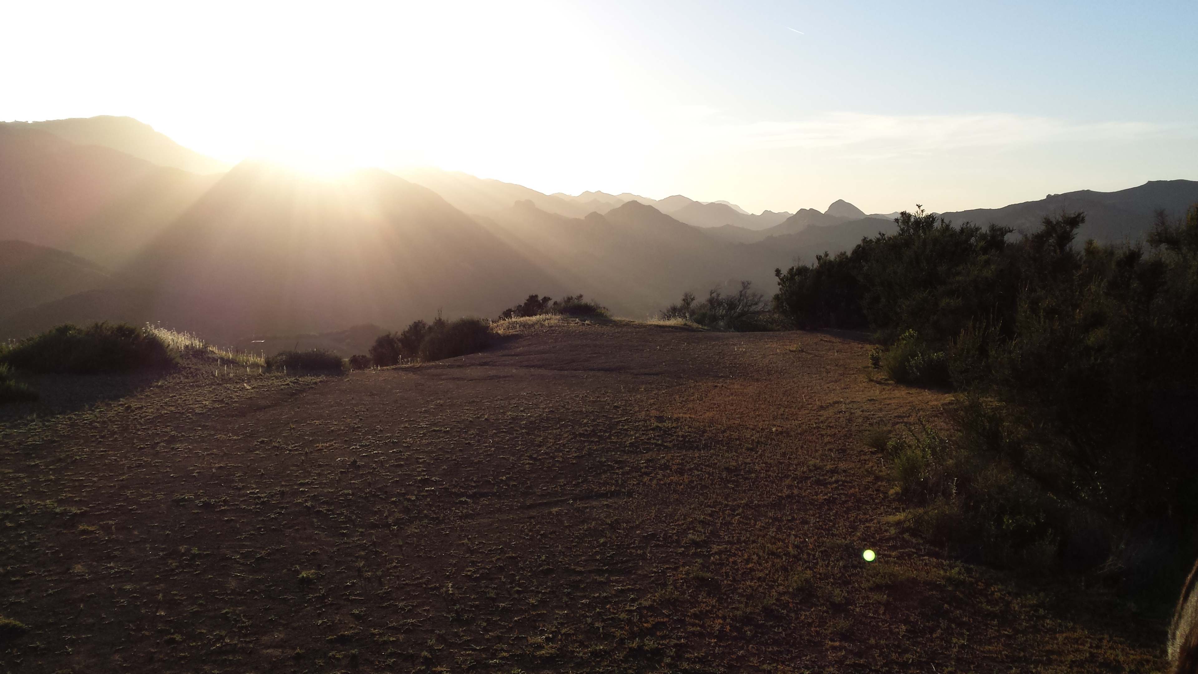 The sun rises over a mountain landscape, casting rays of light across the terrain and creating a silhouette of distant peaks.