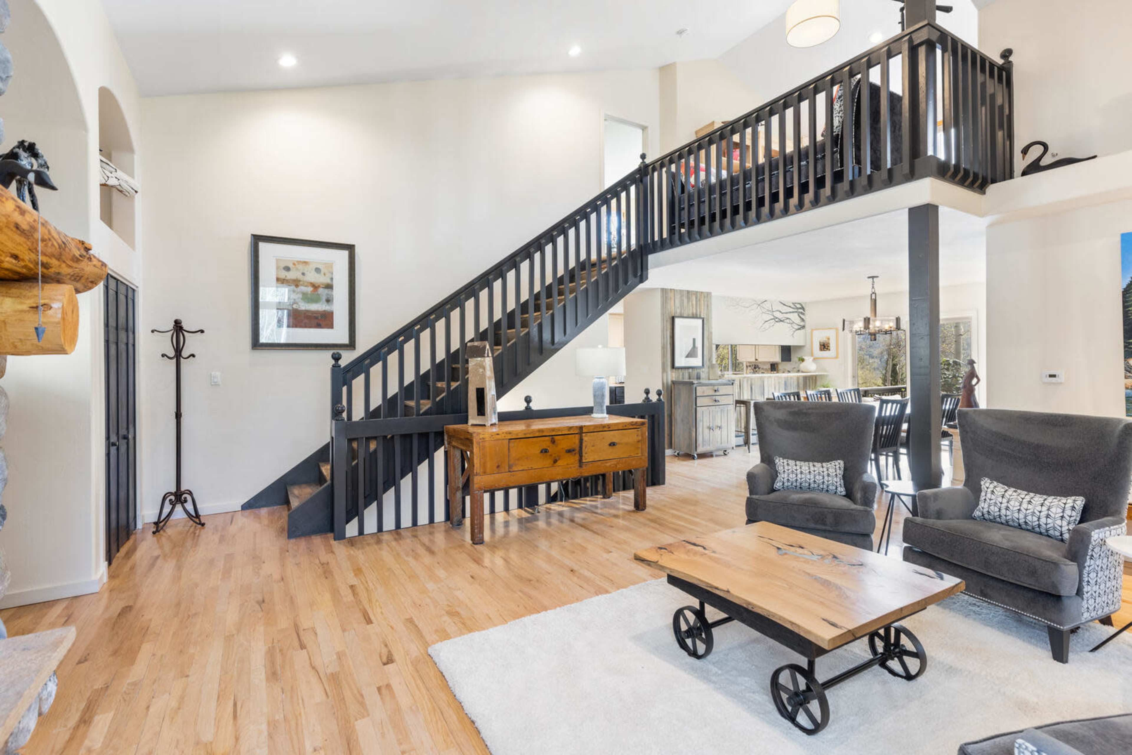 The image shows a spacious living area featuring a staircase, a wooden coffee table on a rug, and two gray armchairs, with a view into a dining area beyond.