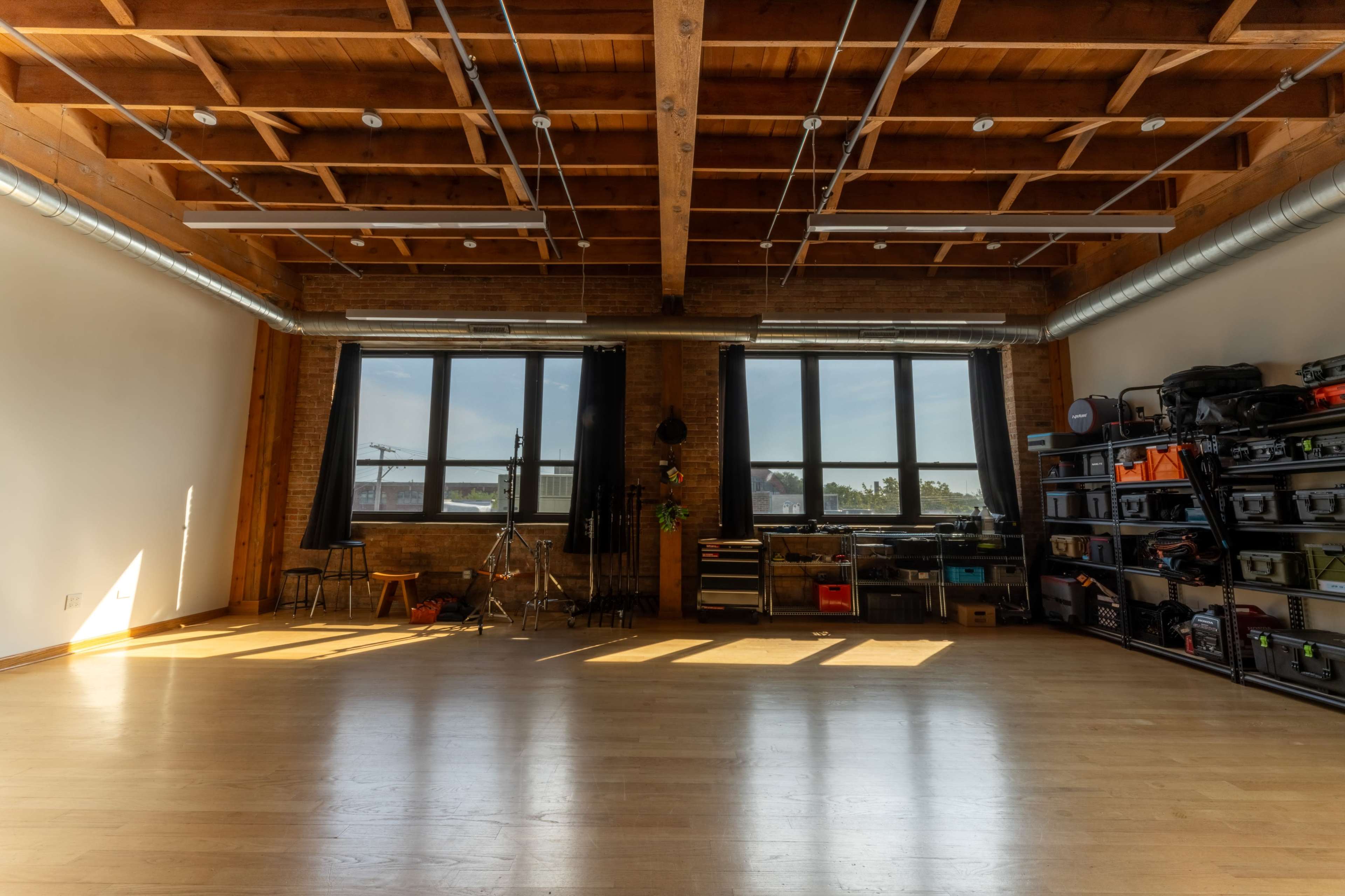 A spacious studio with wooden beams, large windows letting in natural light, and organized storage shelves along one wall.