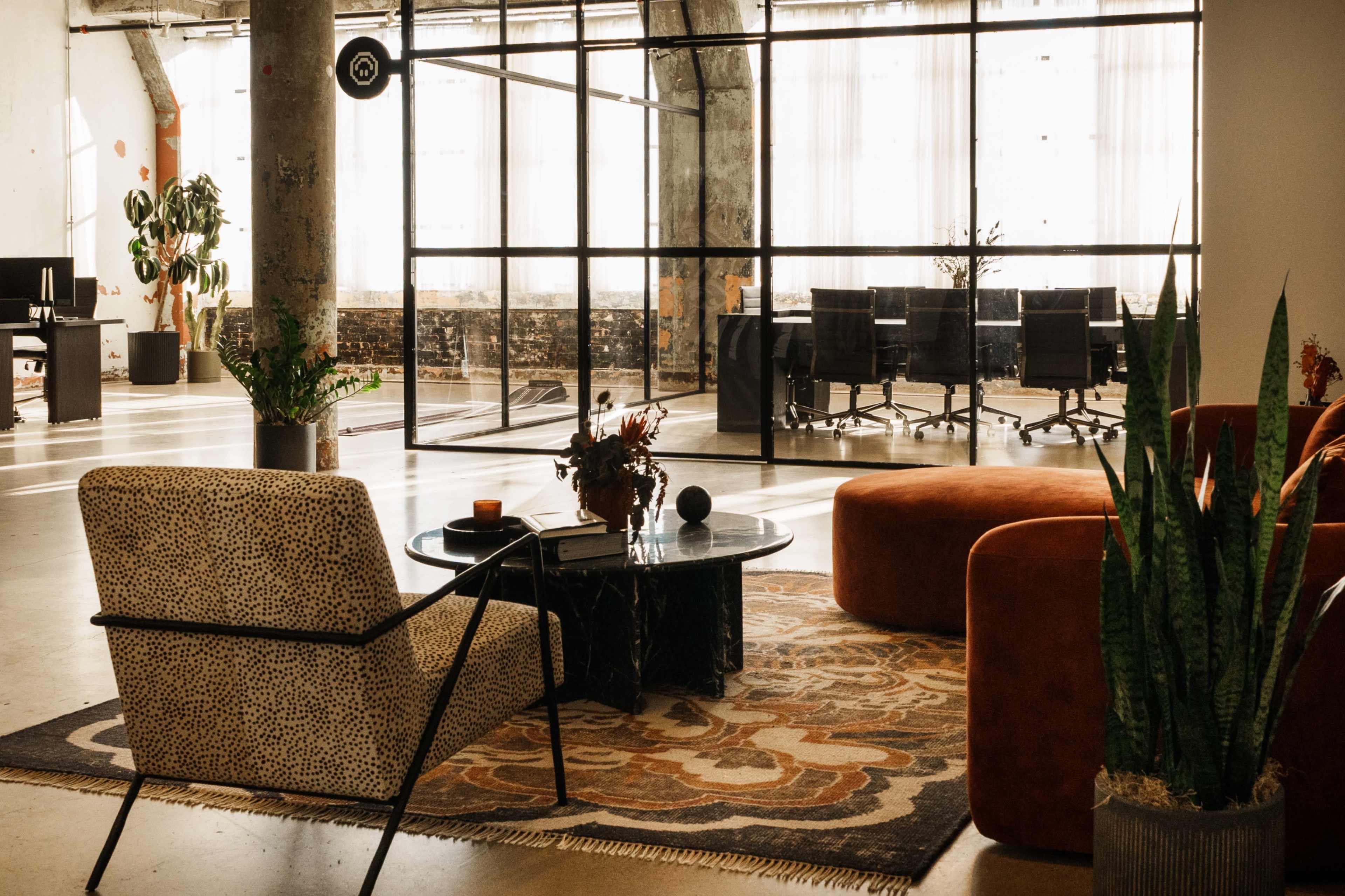 The image shows a modern office space featuring a relaxed seating area with patterned chairs, a round table, and potted plants, alongside a glass-walled conference room visible in the background.