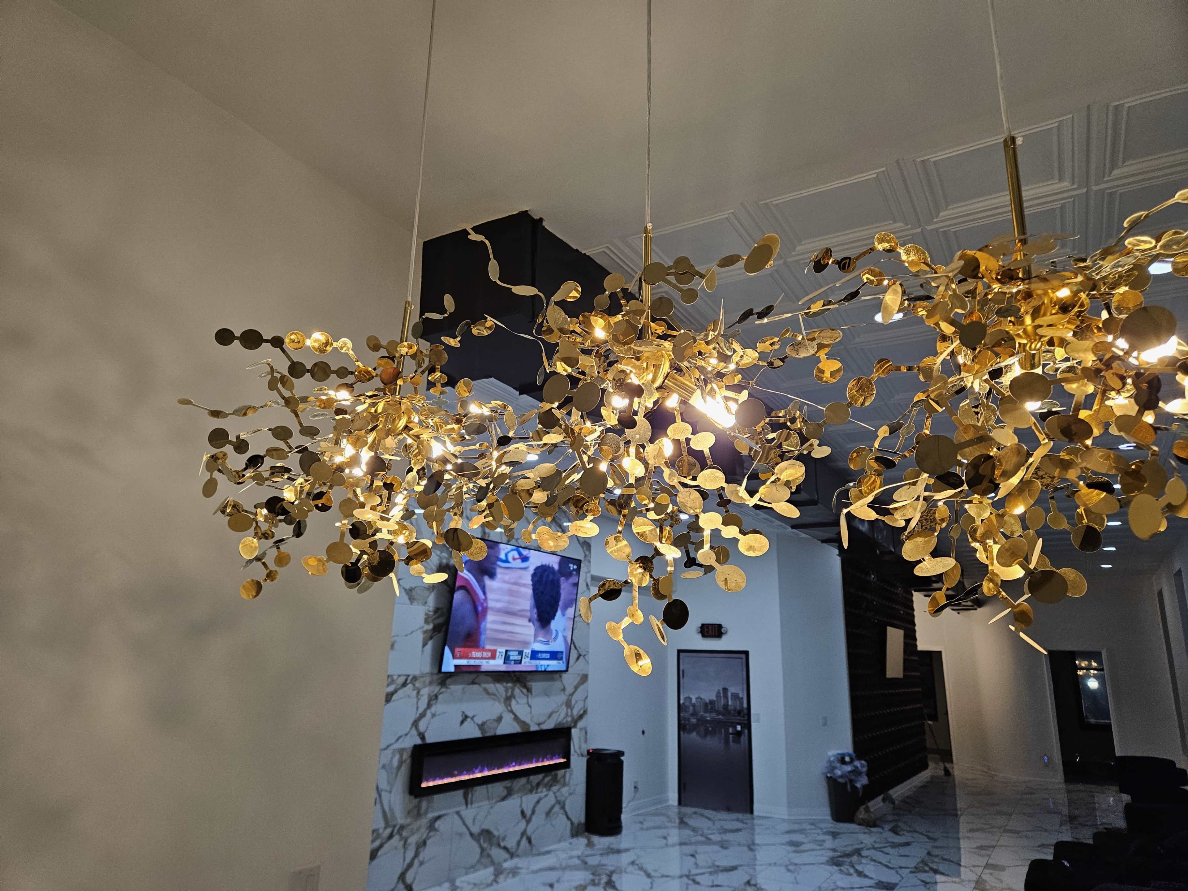 A modern golden chandelier hangs from the ceiling above a marble floor, with a television displaying a program in the background.