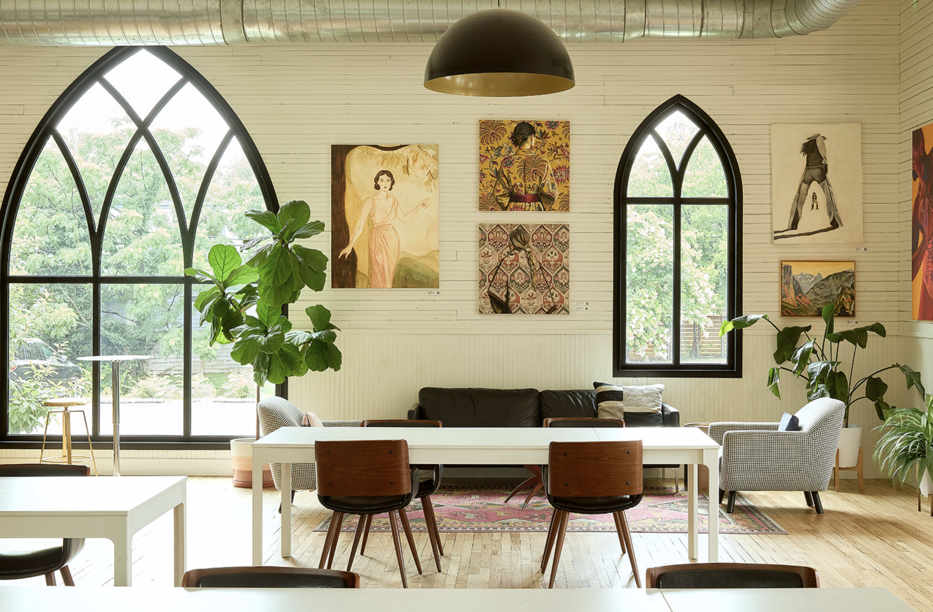 The image shows a bright, modern workspace with large arched windows, plants, and artwork on the walls, featuring a long table and several chairs.