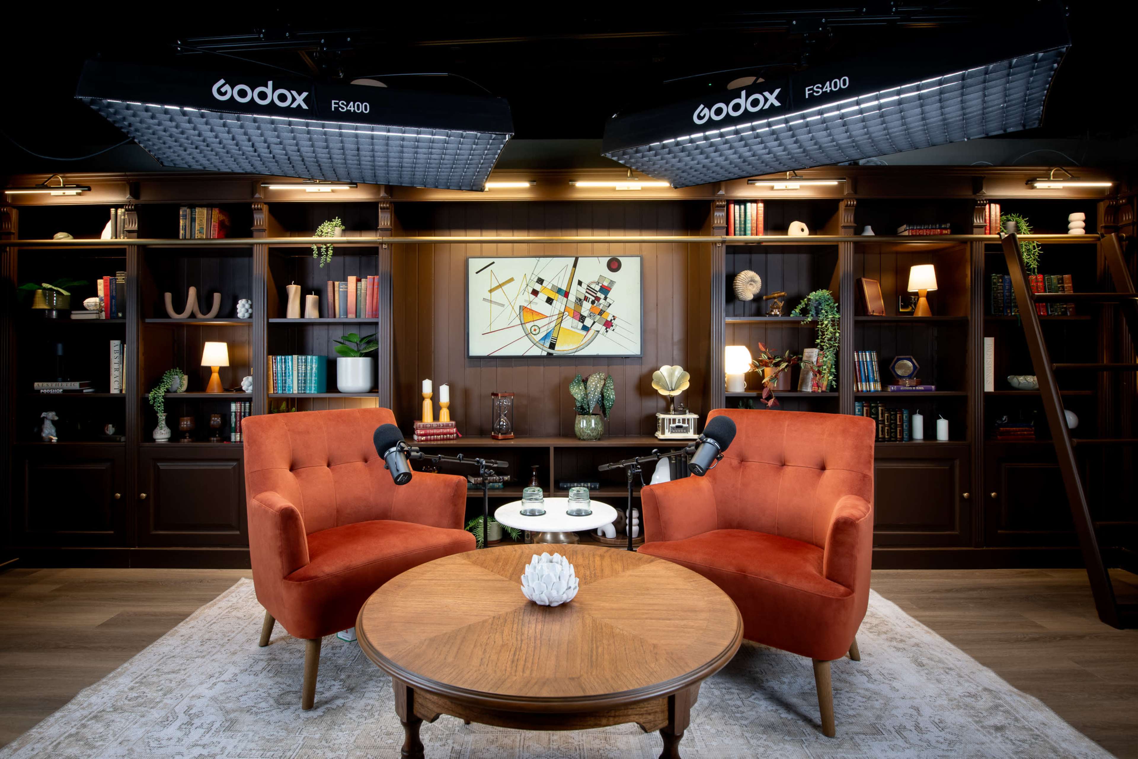 The image shows a cozy studio setup with two orange armchairs facing a wooden table, surrounded by bookshelves filled with books and decorative items, and illuminated by overhead lights.