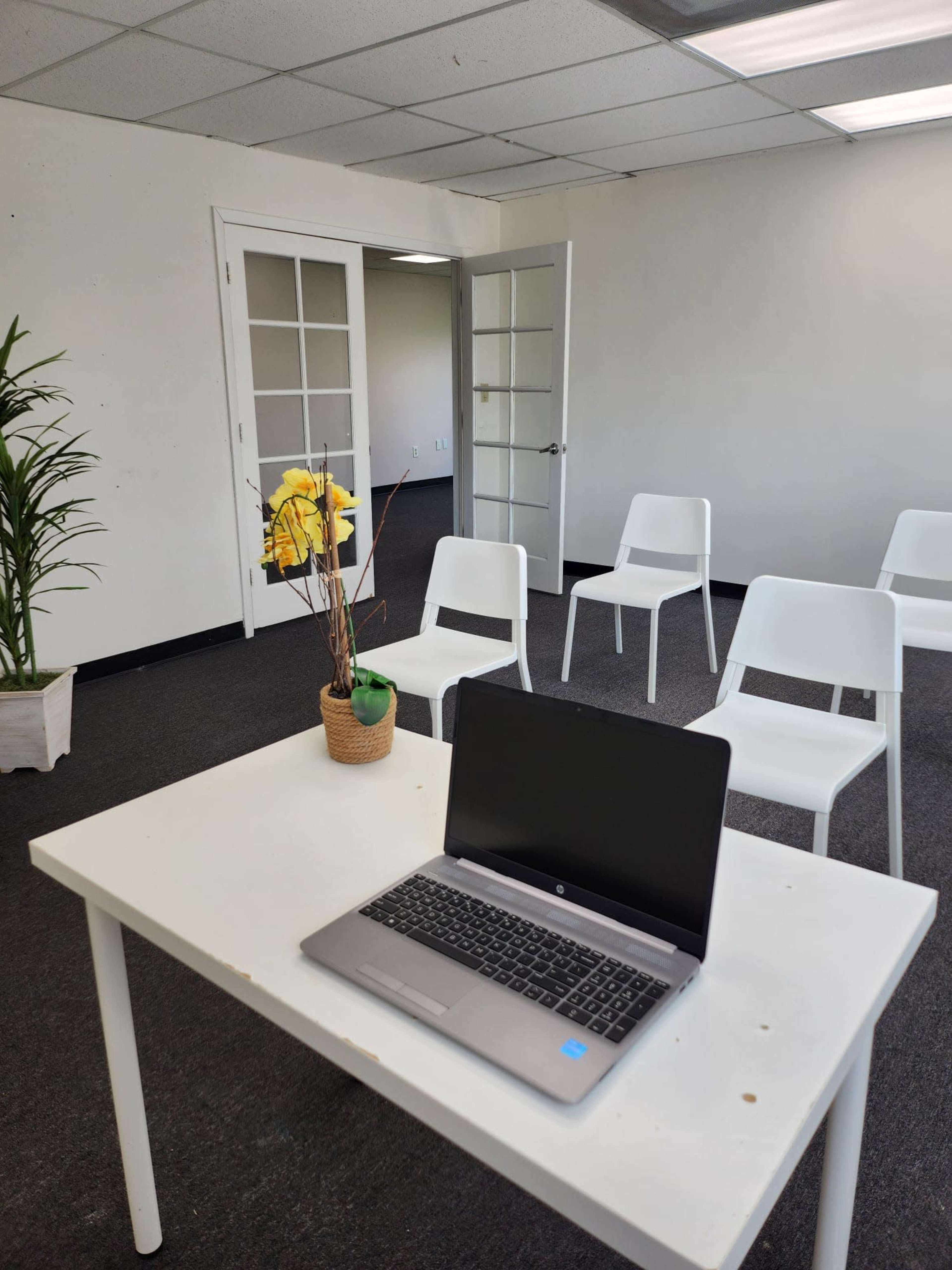 A laptop sits on a table in an empty room with white chairs and a plant in a pot.