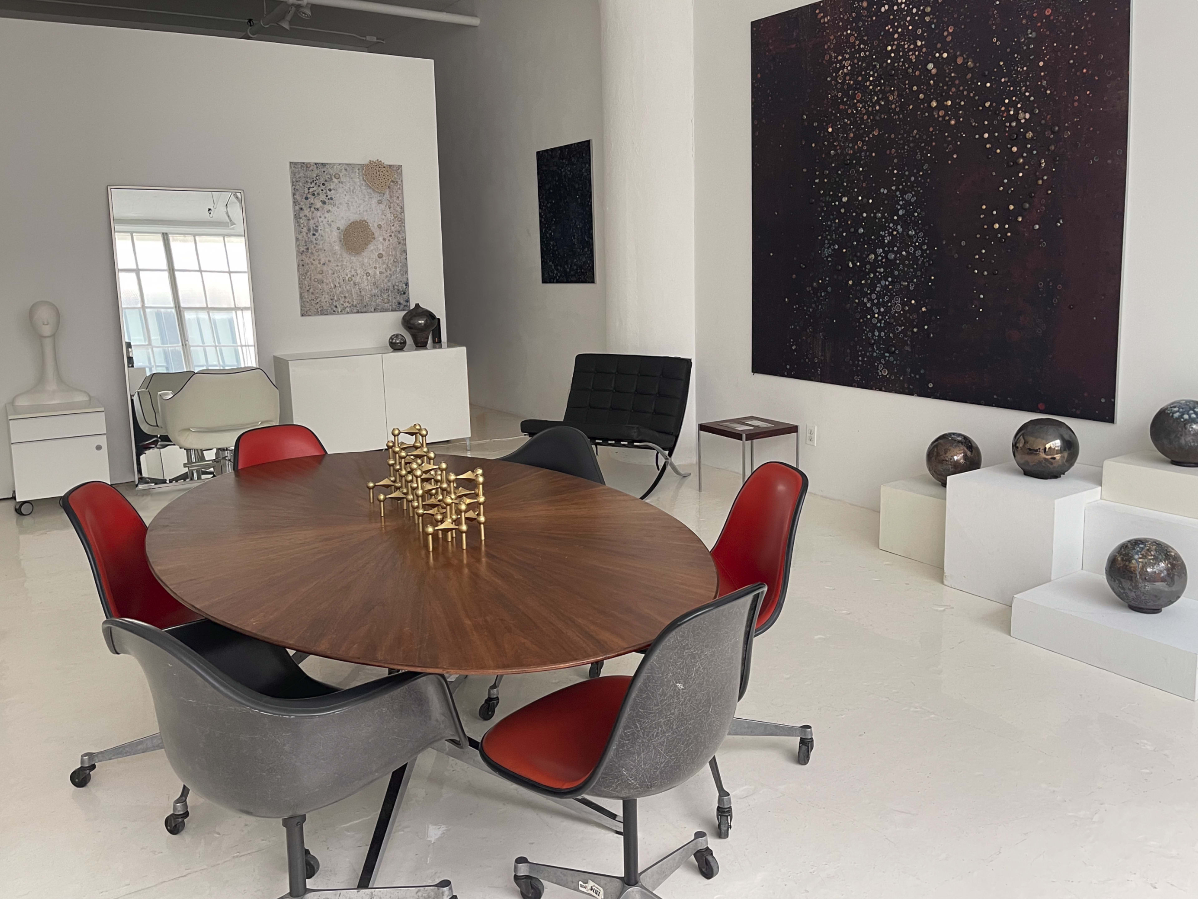 A modern conference room features a round wooden table surrounded by stylish chairs, with abstract artworks and decorative objects displayed on white walls and pedestals.
