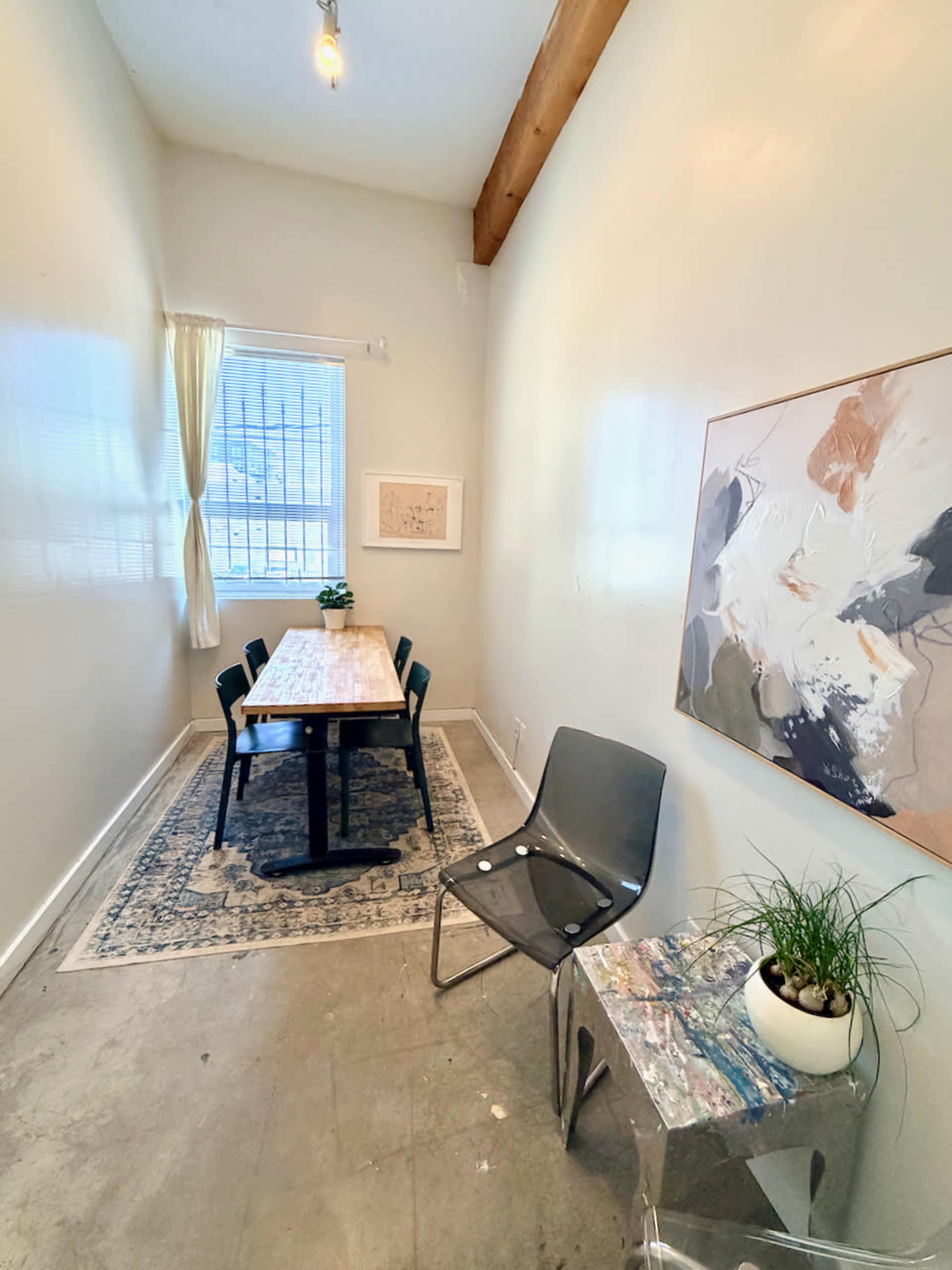 The image shows a narrow, well-lit room with a wooden table and four chairs, adorned with a rug and a decorative painting on the wall.