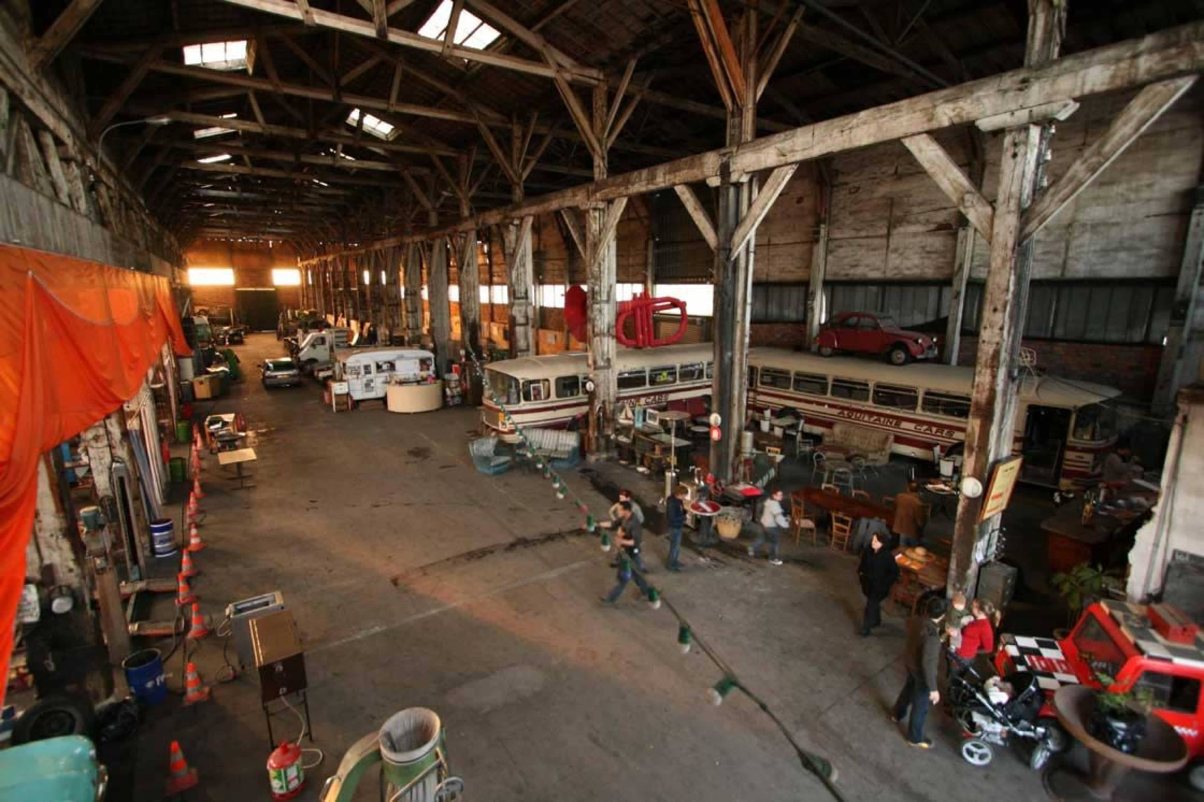The image shows a large, vacant warehouse with vintage vehicles and assorted items scattered throughout the floor space.