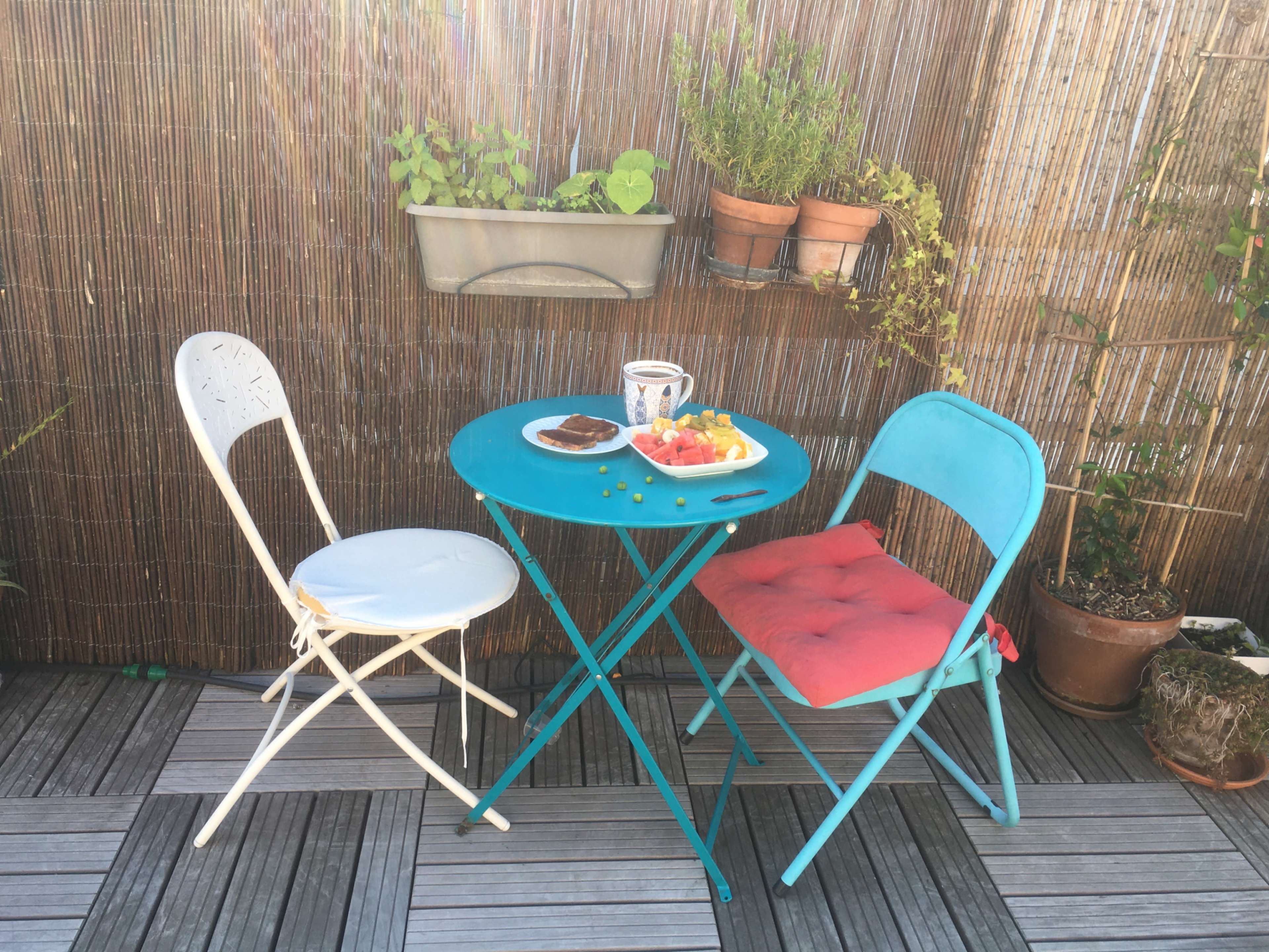 A small blue table with two chairs is set up on a patio, featuring a plate of fruit and a slice of cake.