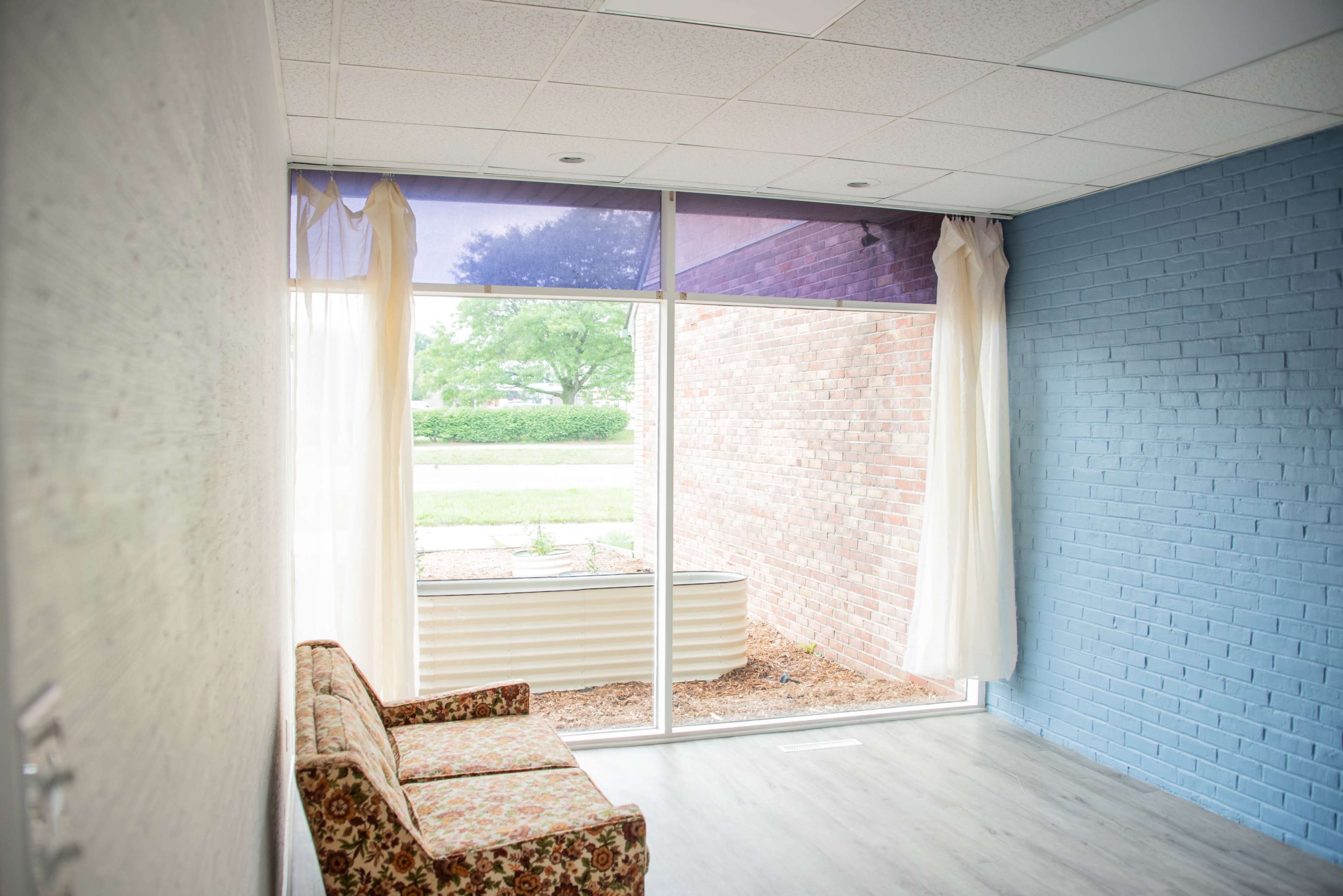 A small room features a floral-patterned couch against one wall, with large windows letting in natural light and revealing a view of an outdoor area.