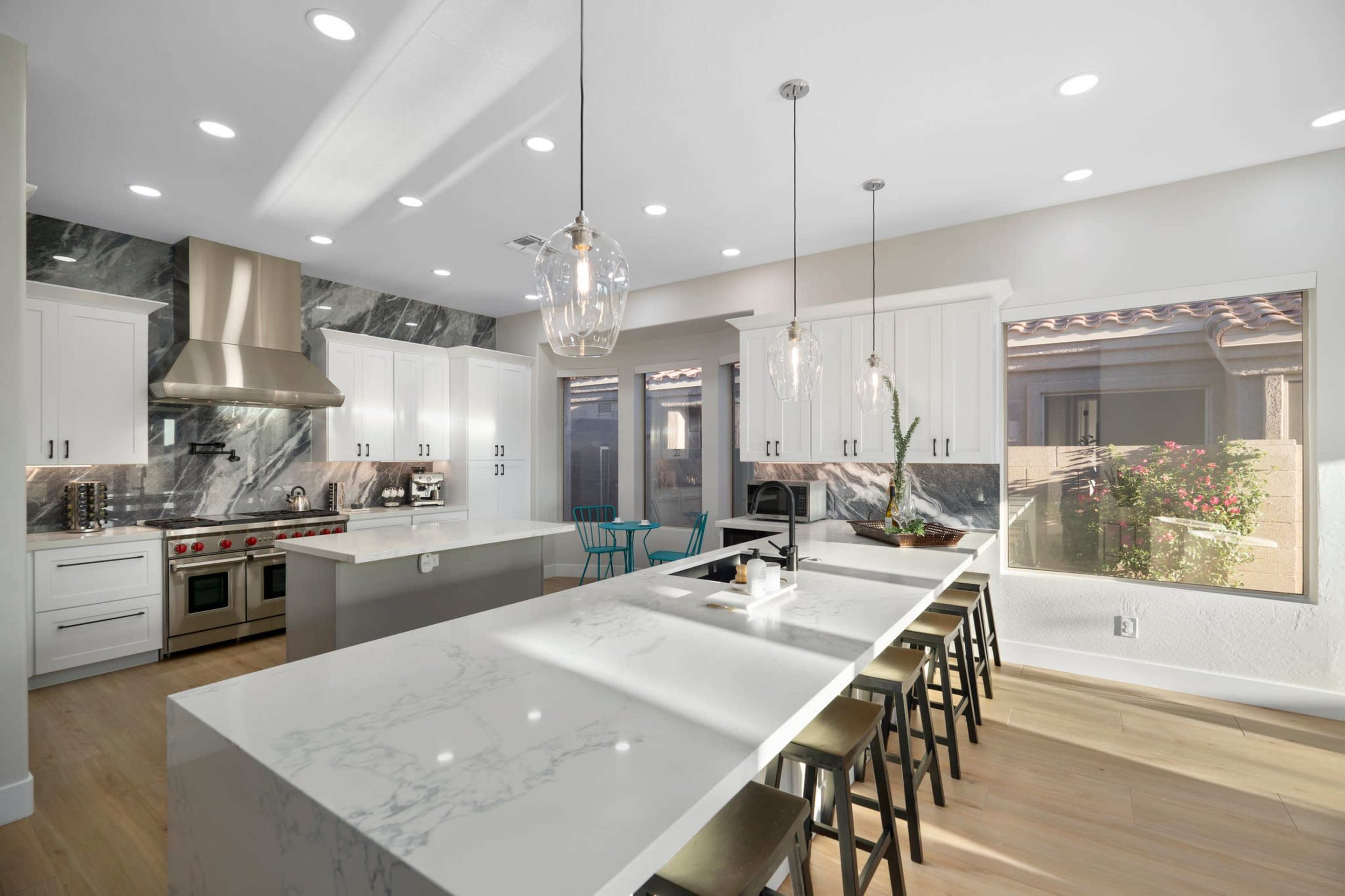 The image shows a modern kitchen featuring white cabinetry, a large marble island with seating, stainless steel appliances, and windows overlooking a landscaped outdoor area.