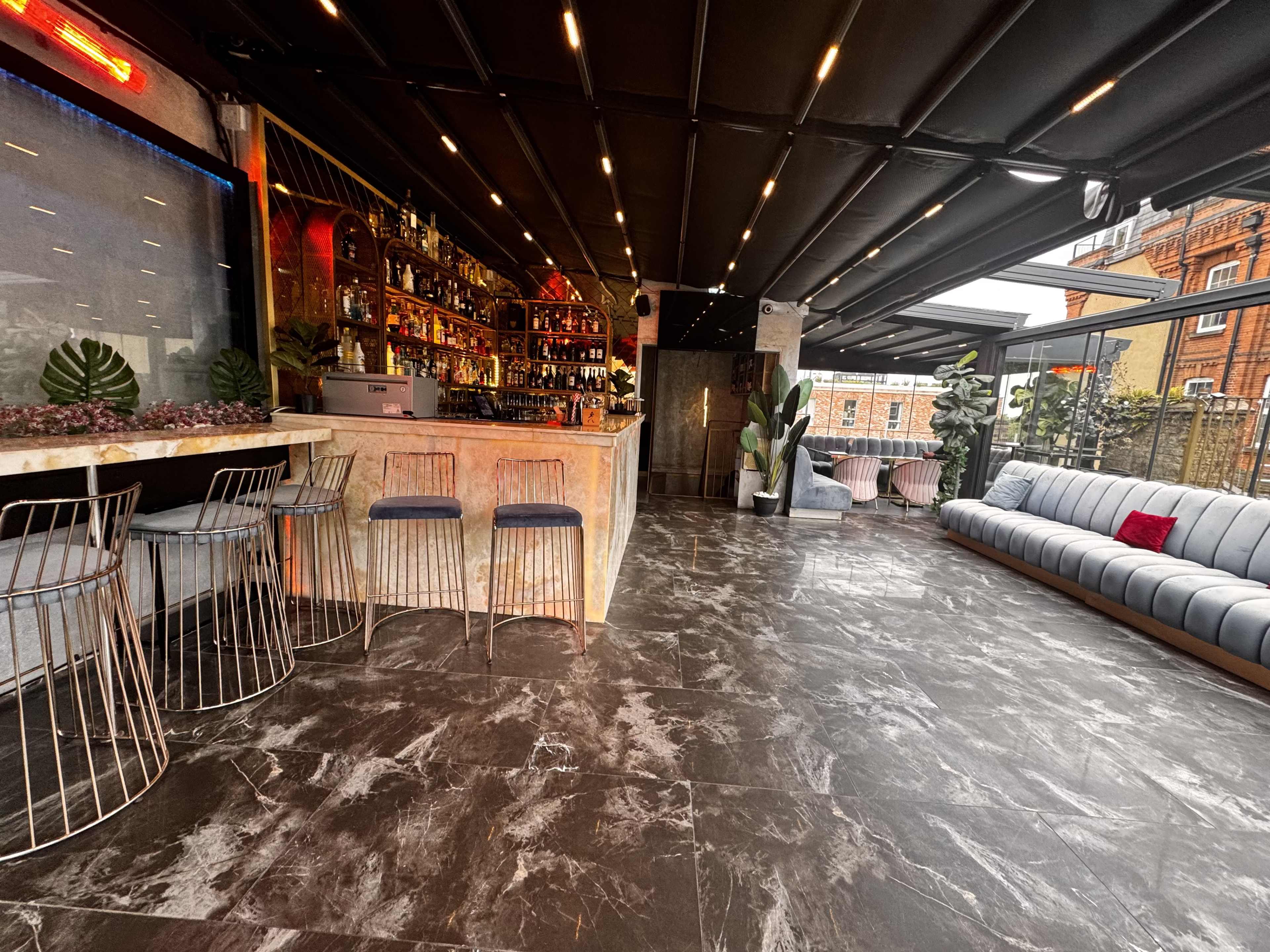 The image shows a modern bar interior with a long counter, high stools, and a spacious seating area featuring a gray couch against large windows.