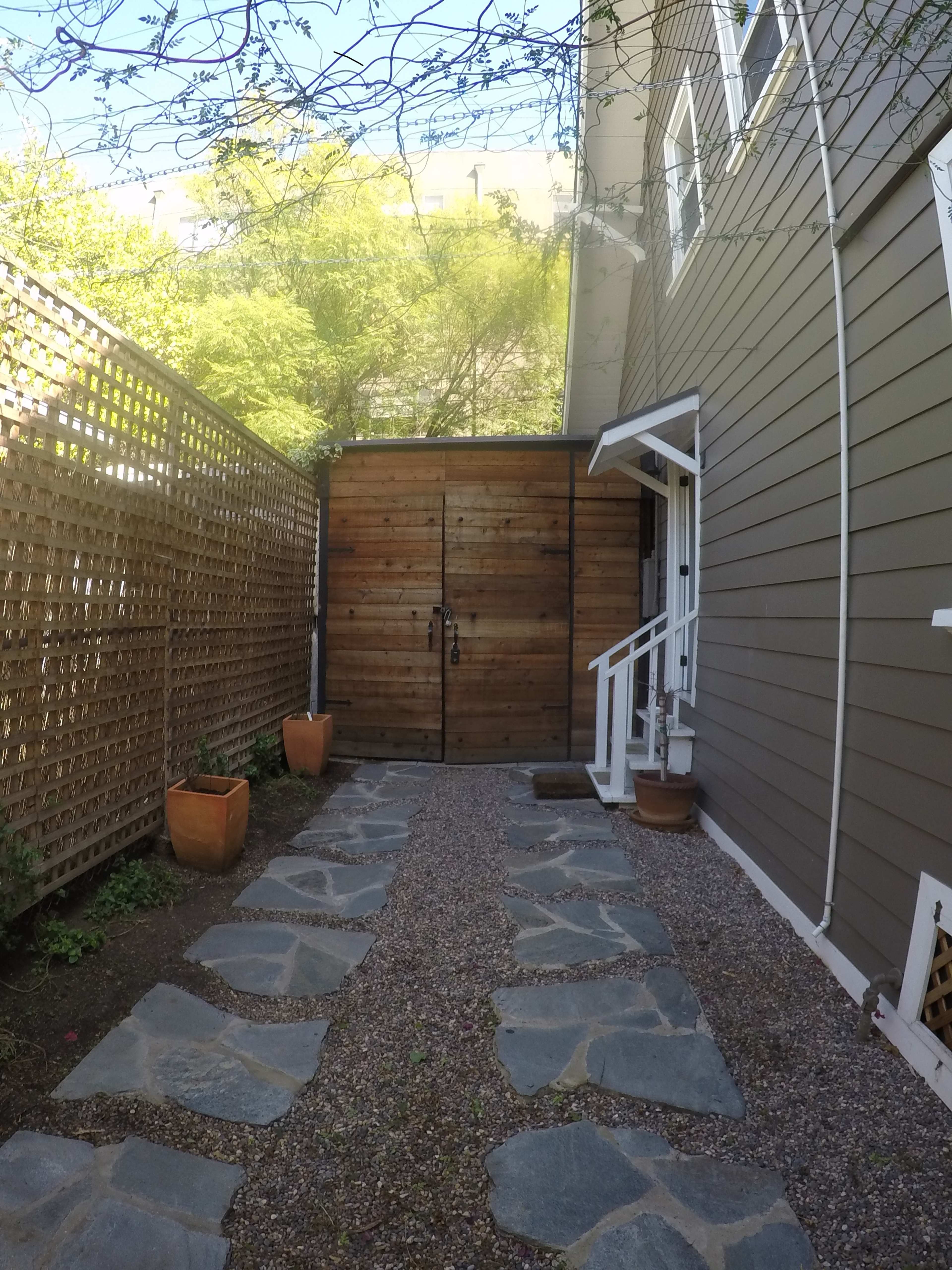 The image shows a pathway made of stone leading to a wooden door bordered by potted plants and surrounded by a wooden lattice fence on one side.