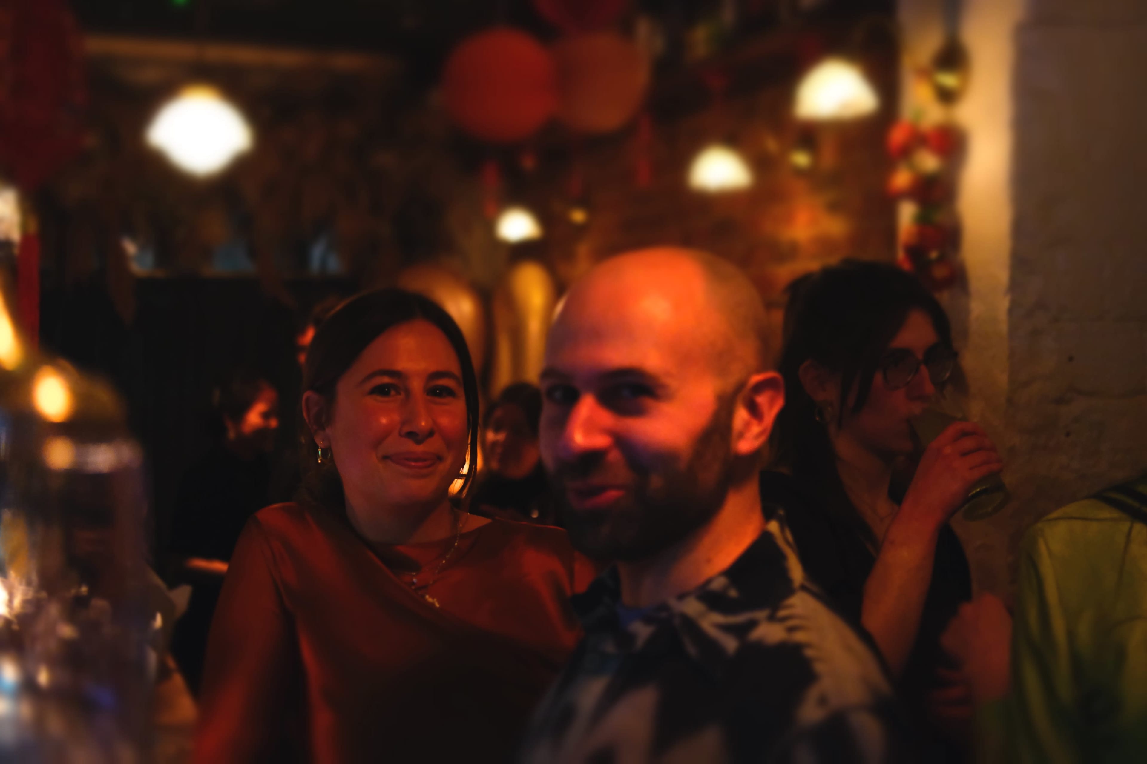 A man and a woman stand close together in a dimly lit setting with soft lighting and decorative elements in the background.