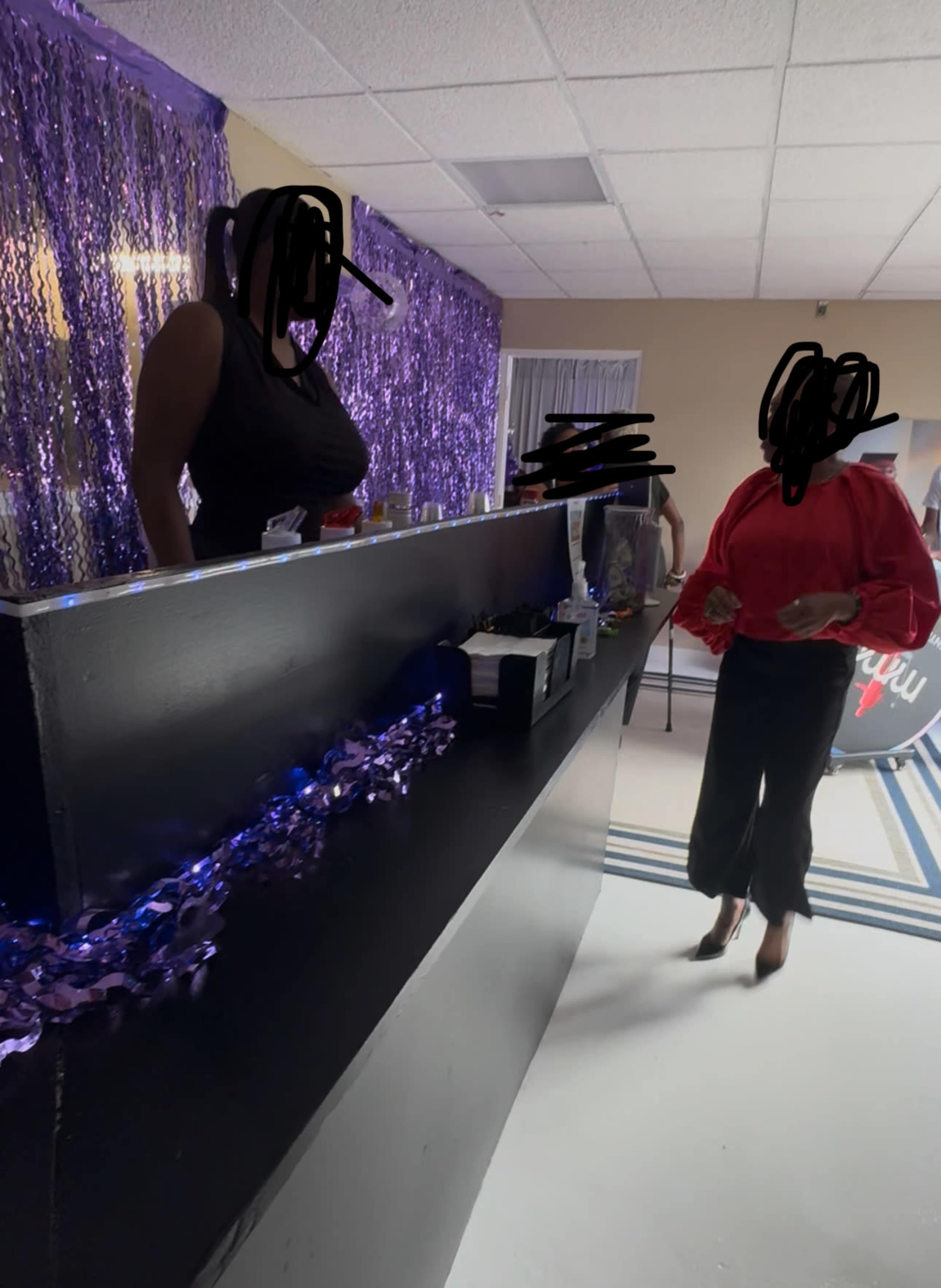 Two individuals stand at a bar adorned with purple decorations, one serving drinks and the other holding a small item, in a brightly lit interior.