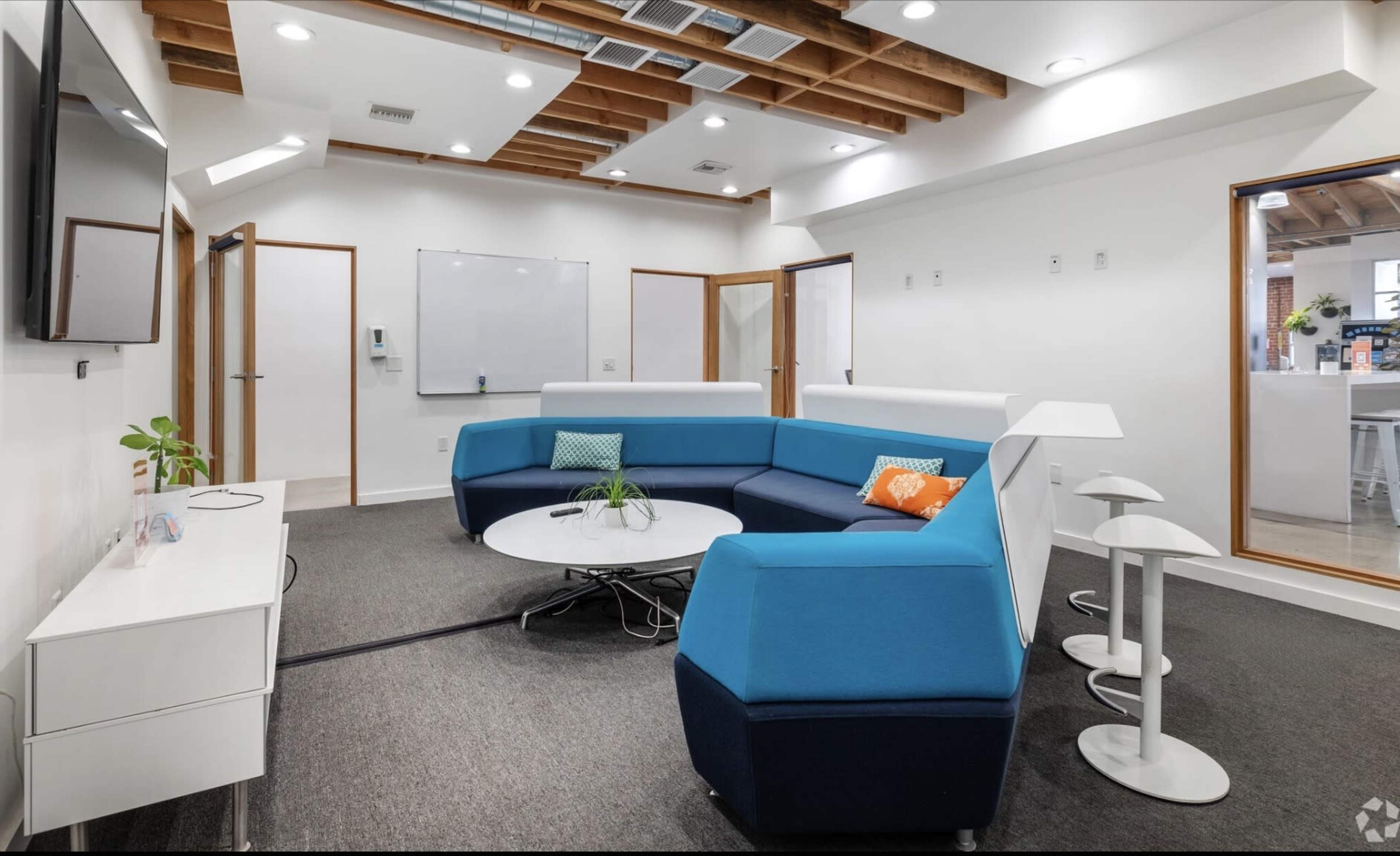 The image shows a modern office space with a blue and white seating area, a round table, and a whiteboard on a wall.