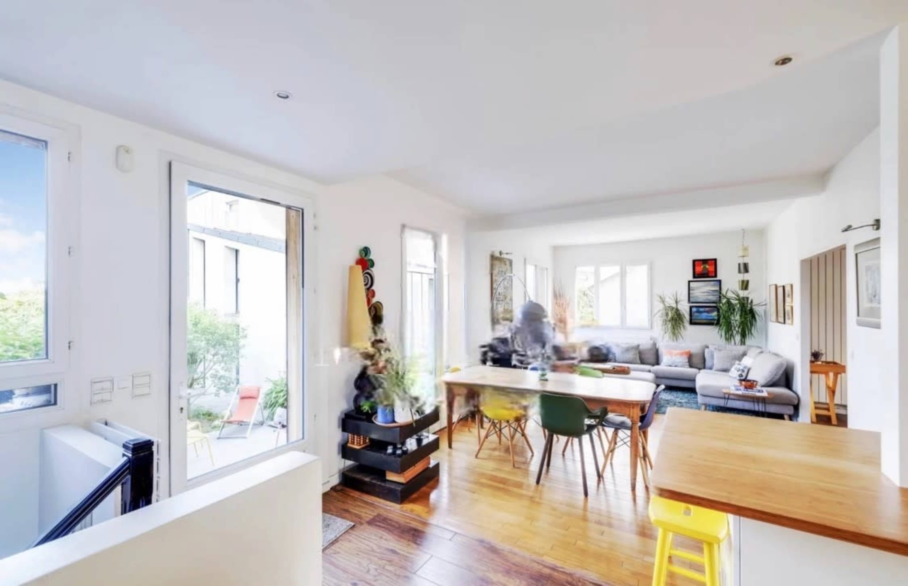 A bright, modern living space features a sofa, dining area, and large windows with plants and artwork.