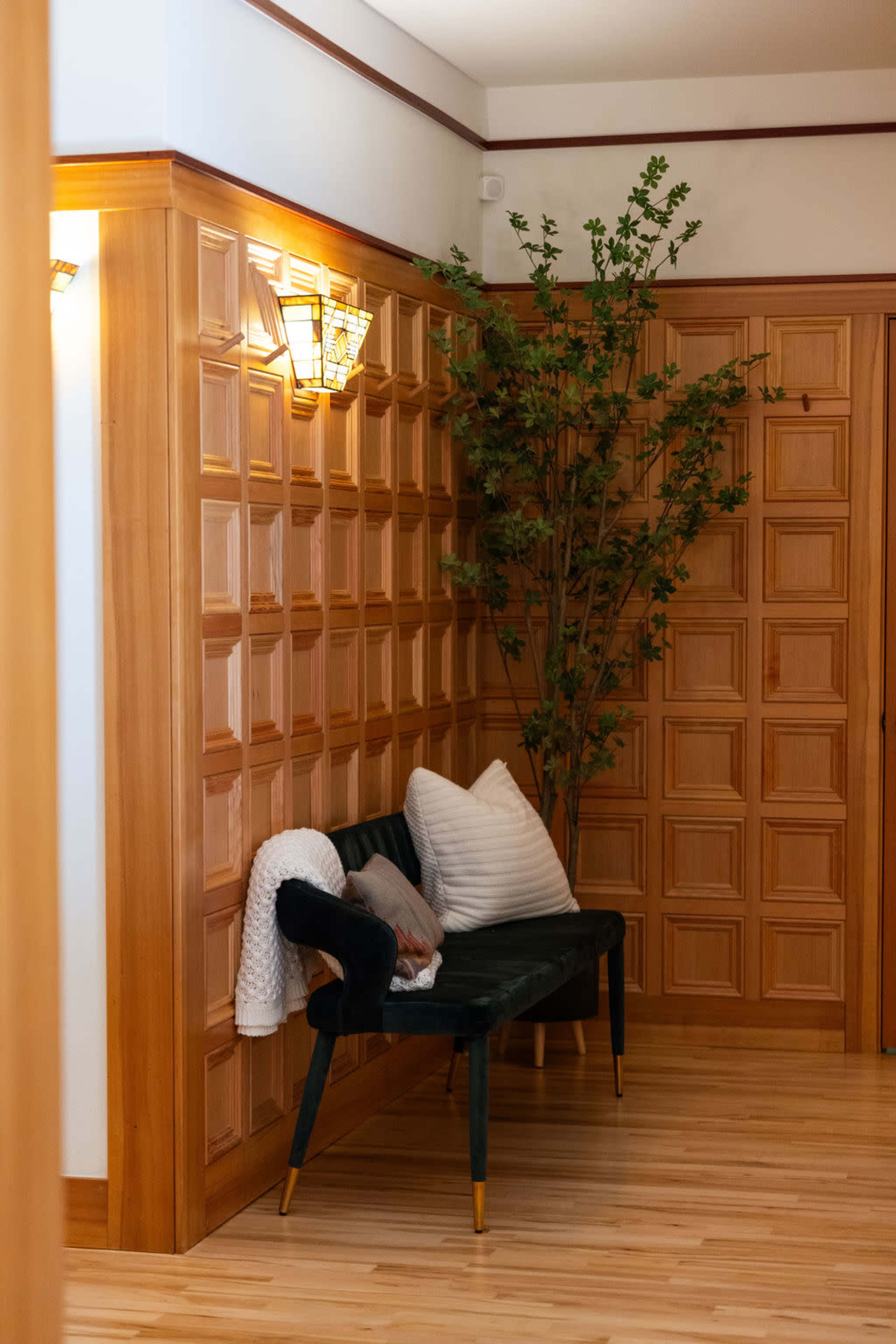 A cozy entryway featuring a dark green bench with cushions, a tall potted plant, and paneled wooden walls illuminated by wall sconces.