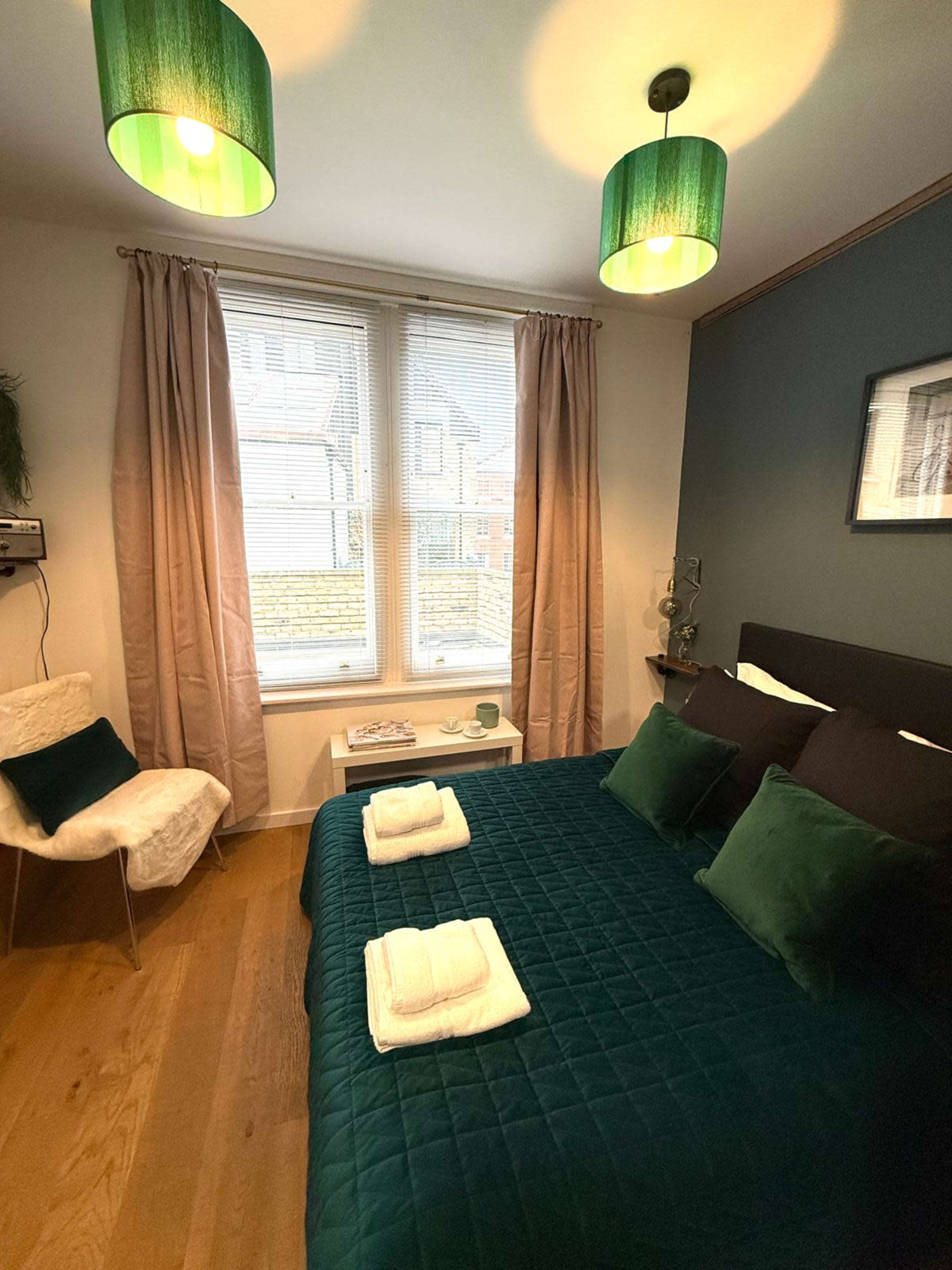 The image shows a cozy bedroom featuring a green quilted bedspread, two white towels neatly arranged on the bed, a small bedside table, and a chair by a window with light curtains.