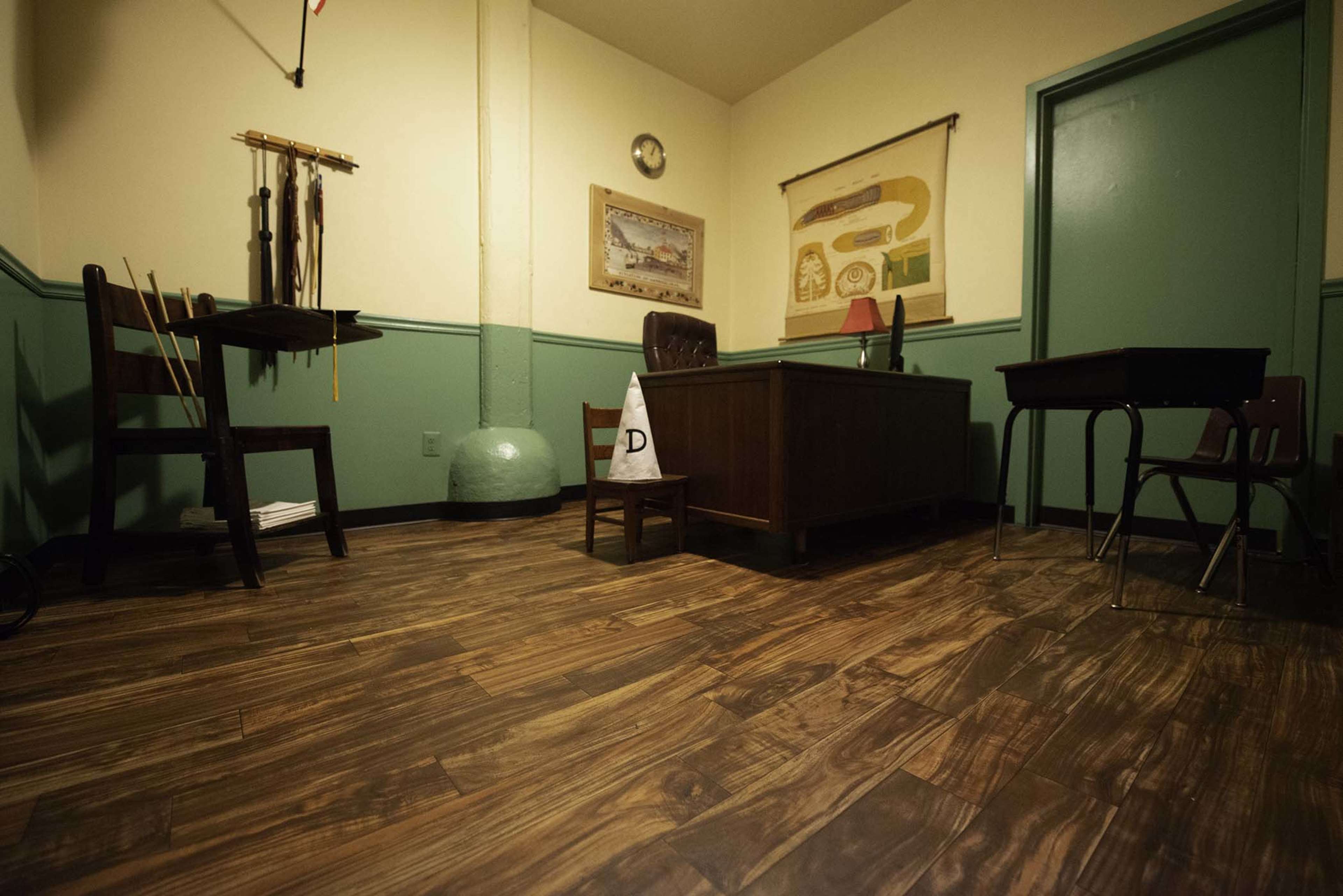 The image shows a sparsely furnished room with a desk, chairs, and a poster on the wall.