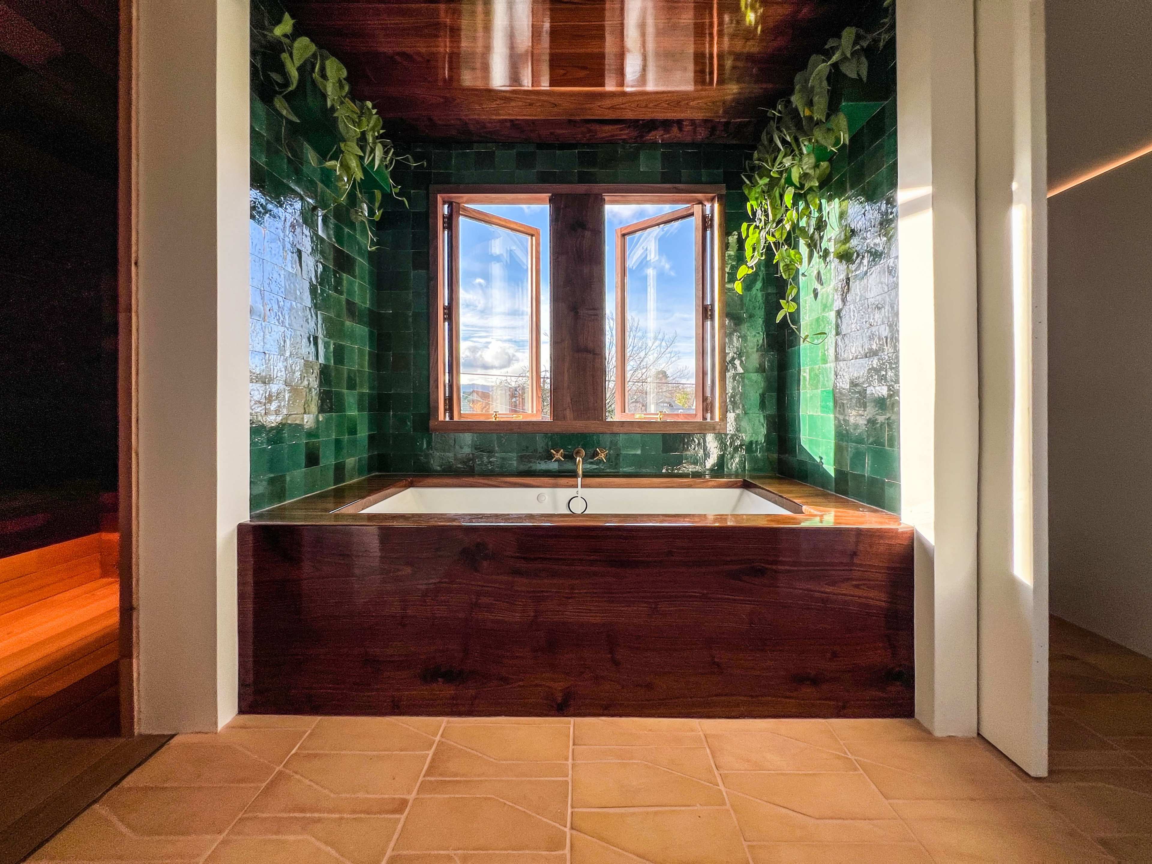 The image shows a modern bathroom with a green-tiled wall, a wooden bathtub, and two windows that provide natural light.
