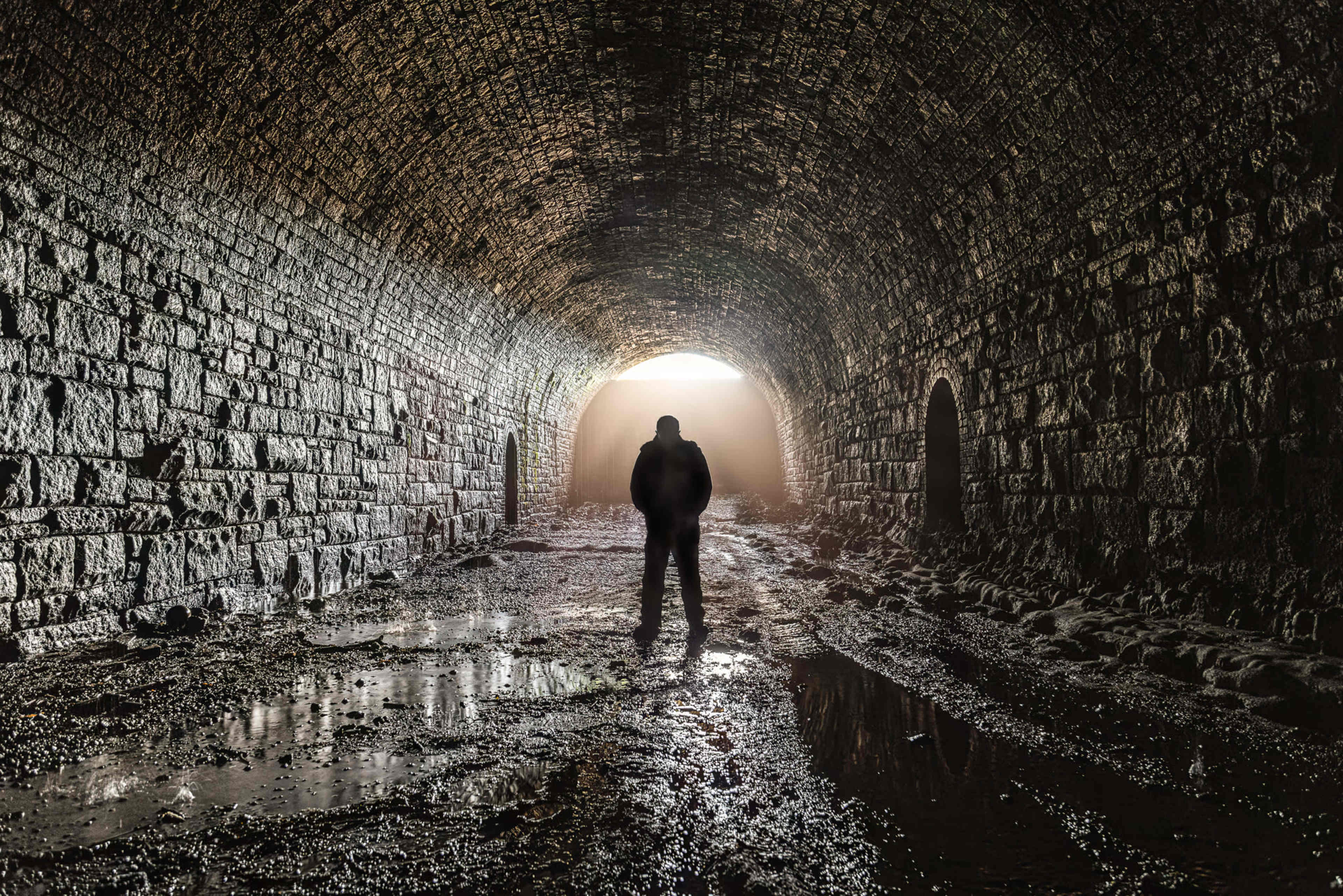 A person stands in a dimly lit stone tunnel, with light streaming in from the entrance and water pooling on the ground.