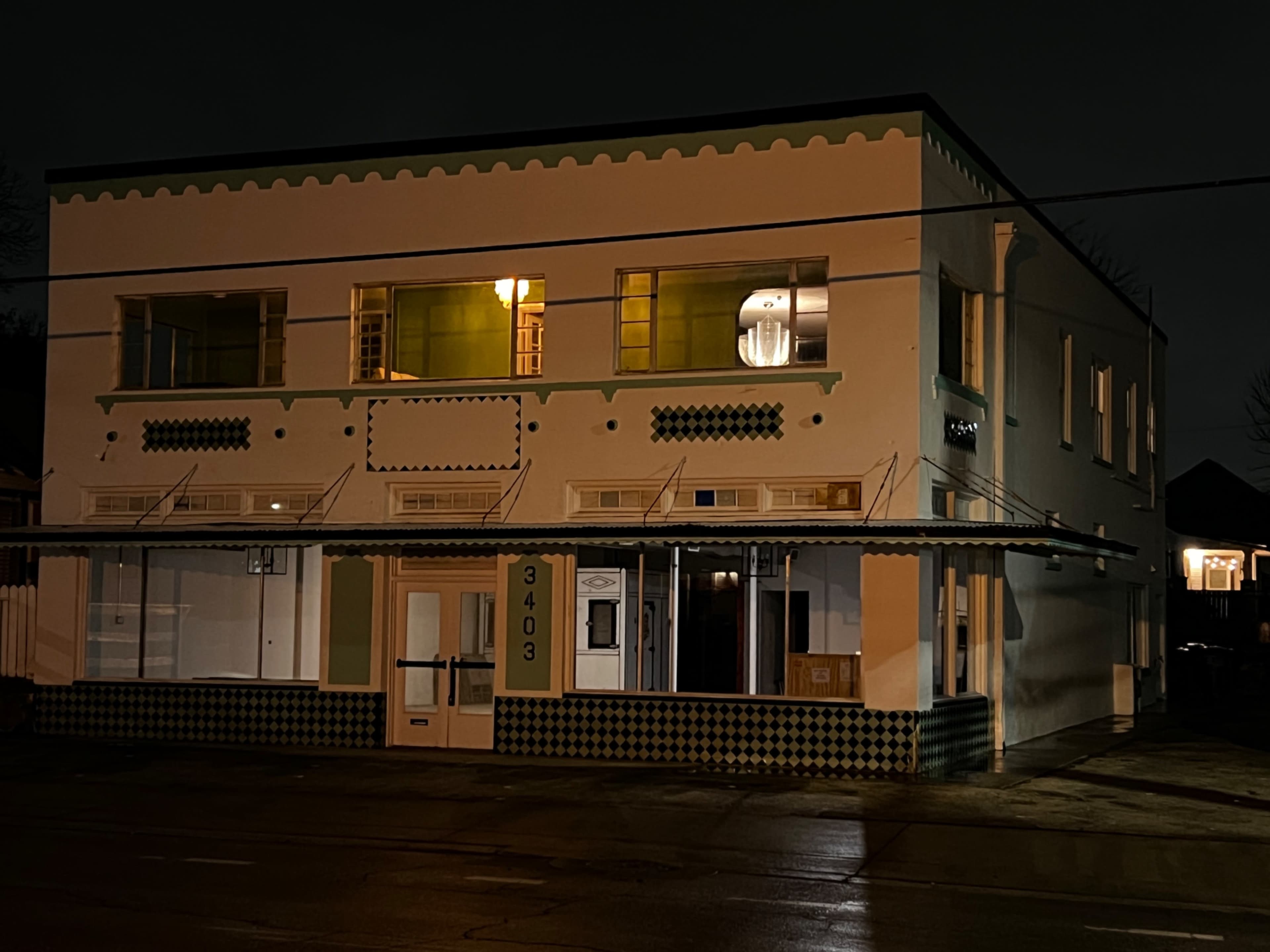 The image shows a two-story, brightly lit building with a patterned exterior and storefront on a dark, wet street at night.