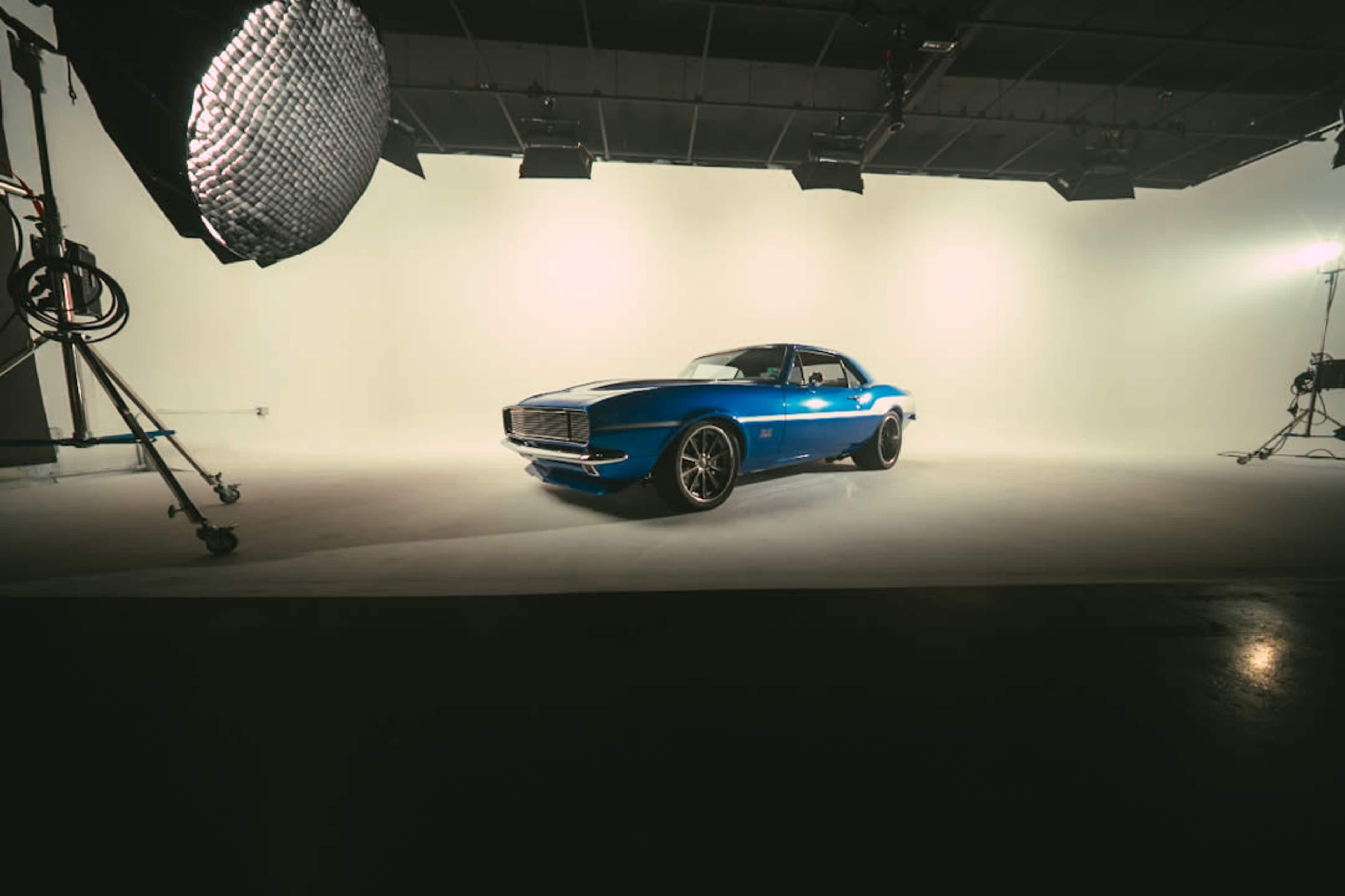 A blue vintage car is positioned in a well-lit studio against a plain white backdrop.