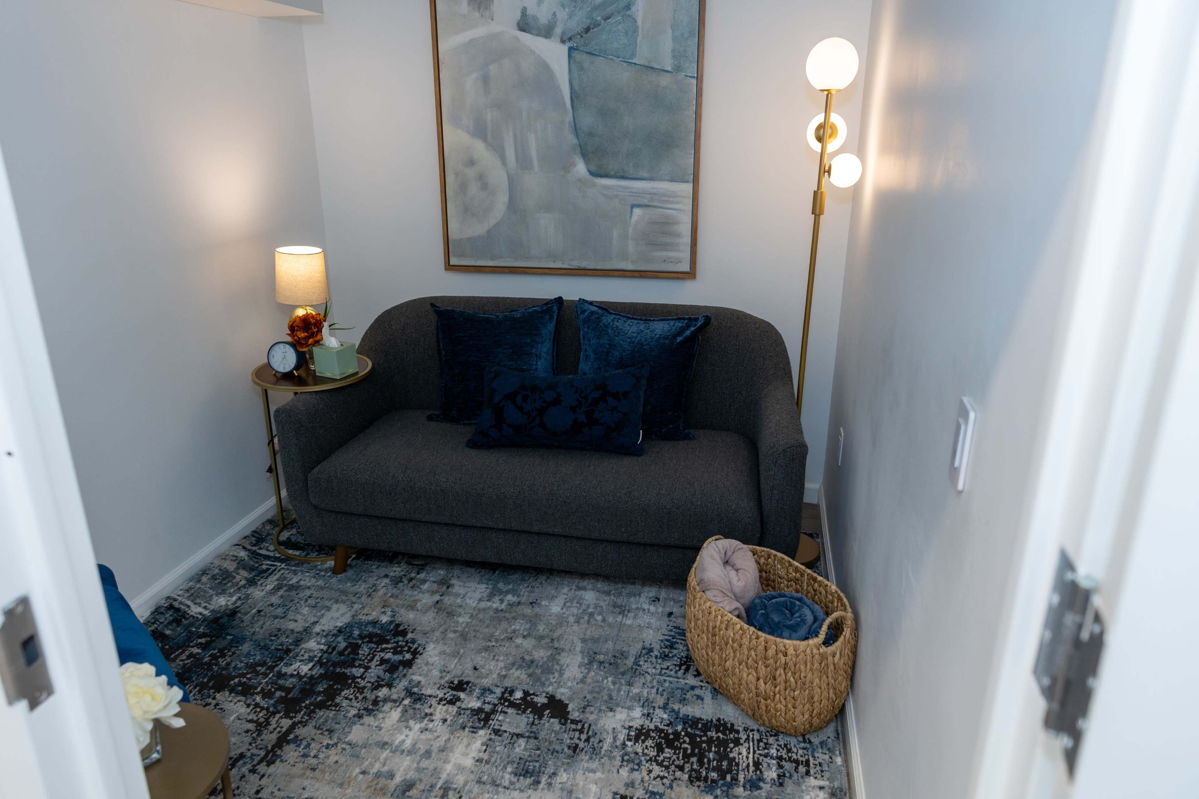 The image shows a cozy corner of a room featuring a gray sofa with blue pillows, a small side table with a lamp, a wall art piece, and a woven basket on a patterned rug.
