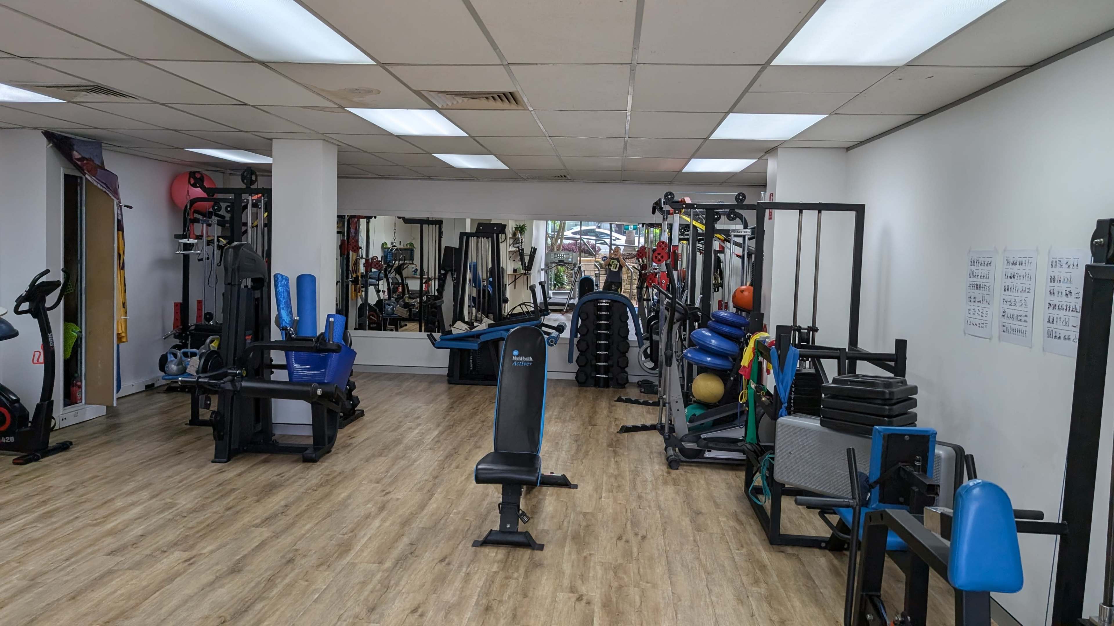 The image depicts an indoor gym with various workout equipment arranged neatly, including weight machines, fitness balls, and exercise mats.