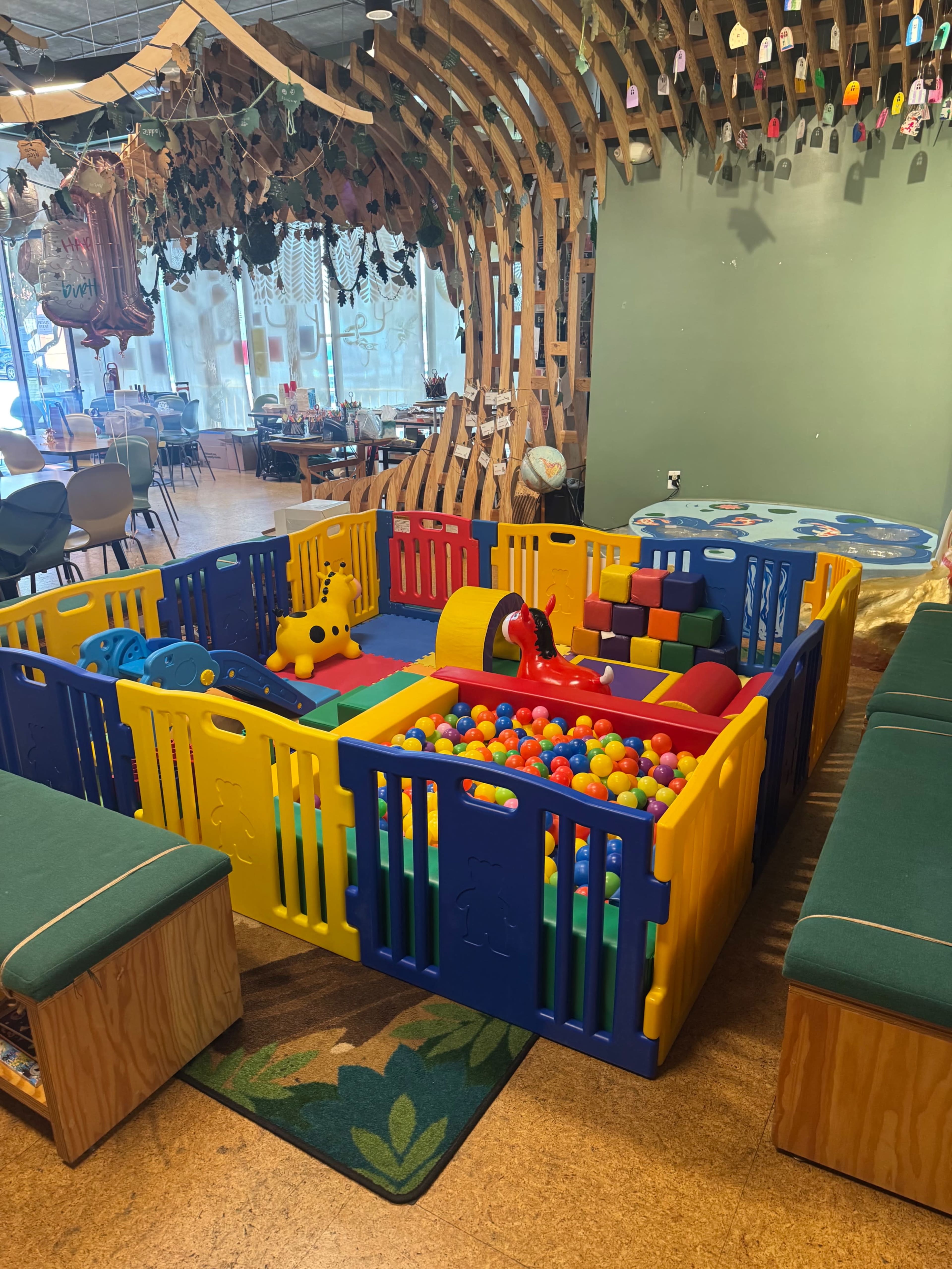 The image shows a colorful children’s playpen filled with various soft toys and balls, surrounded by seating areas in a play space designed for kids.