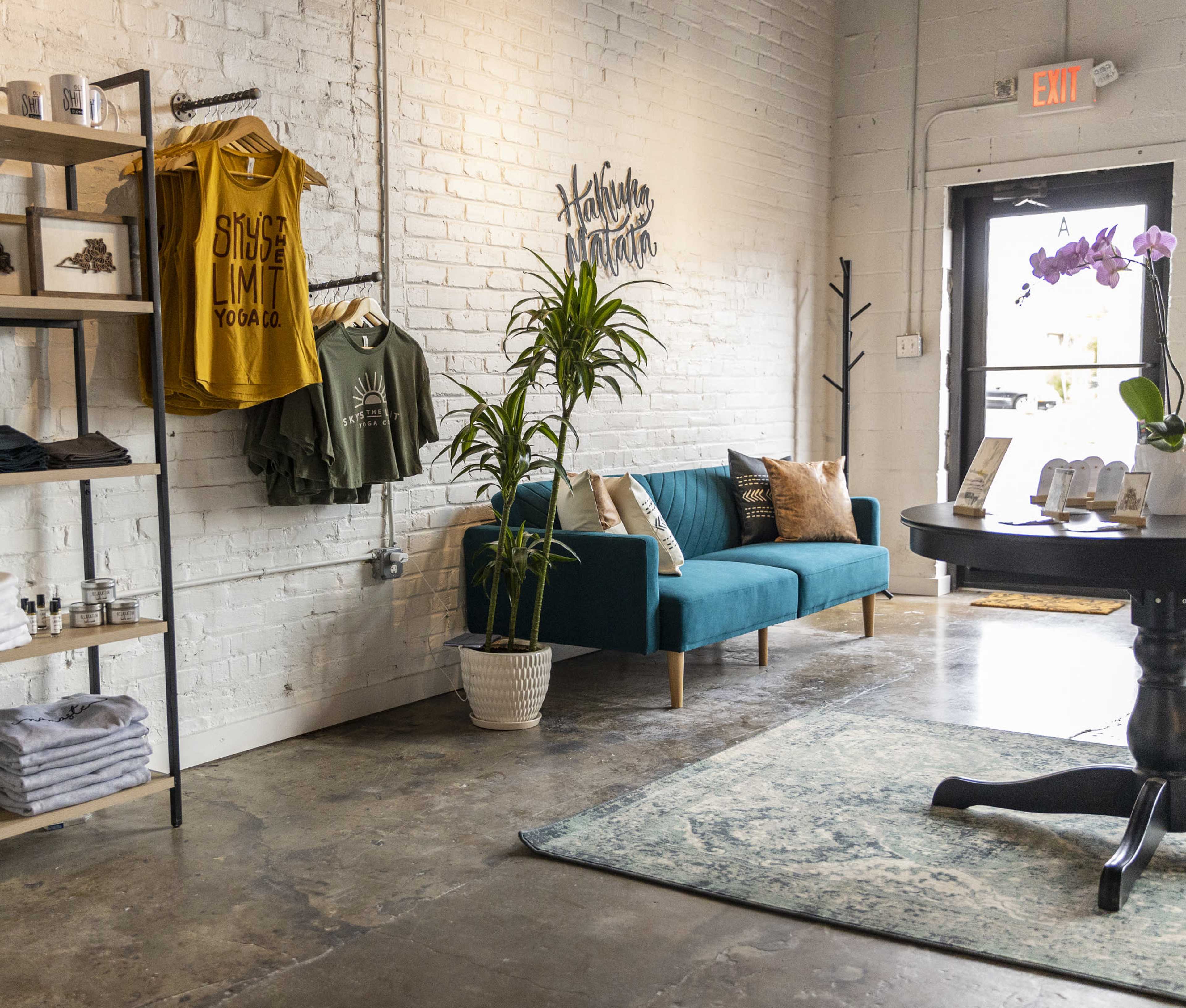 A retail space featuring a turquoise couch, a display of clothing on a wooden rack, and a table with brochures and a plant near the entrance.