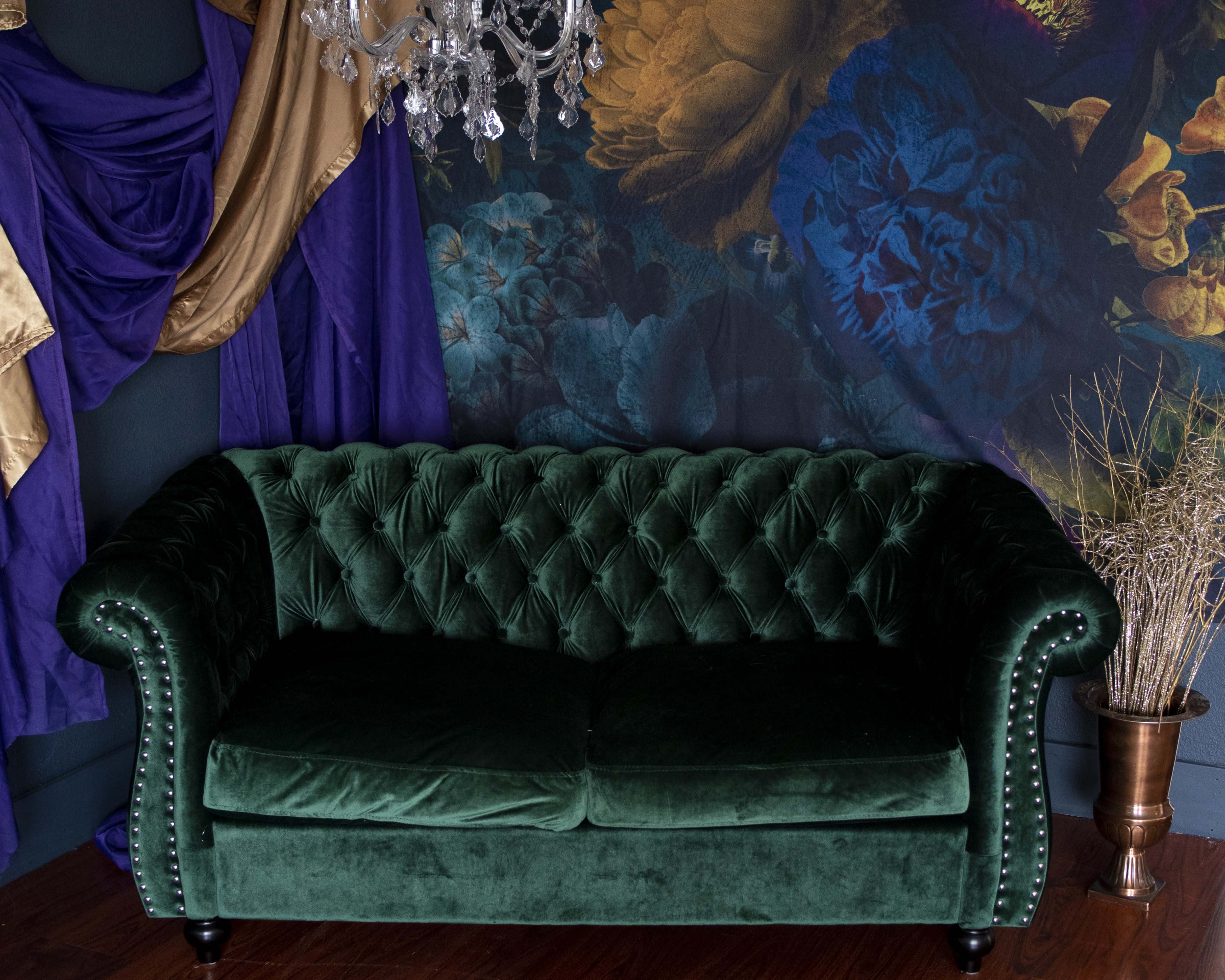 The image features a green velvet tufted sofa in front of a colorful floral wall tapestry, with draped fabric in gold and purple.