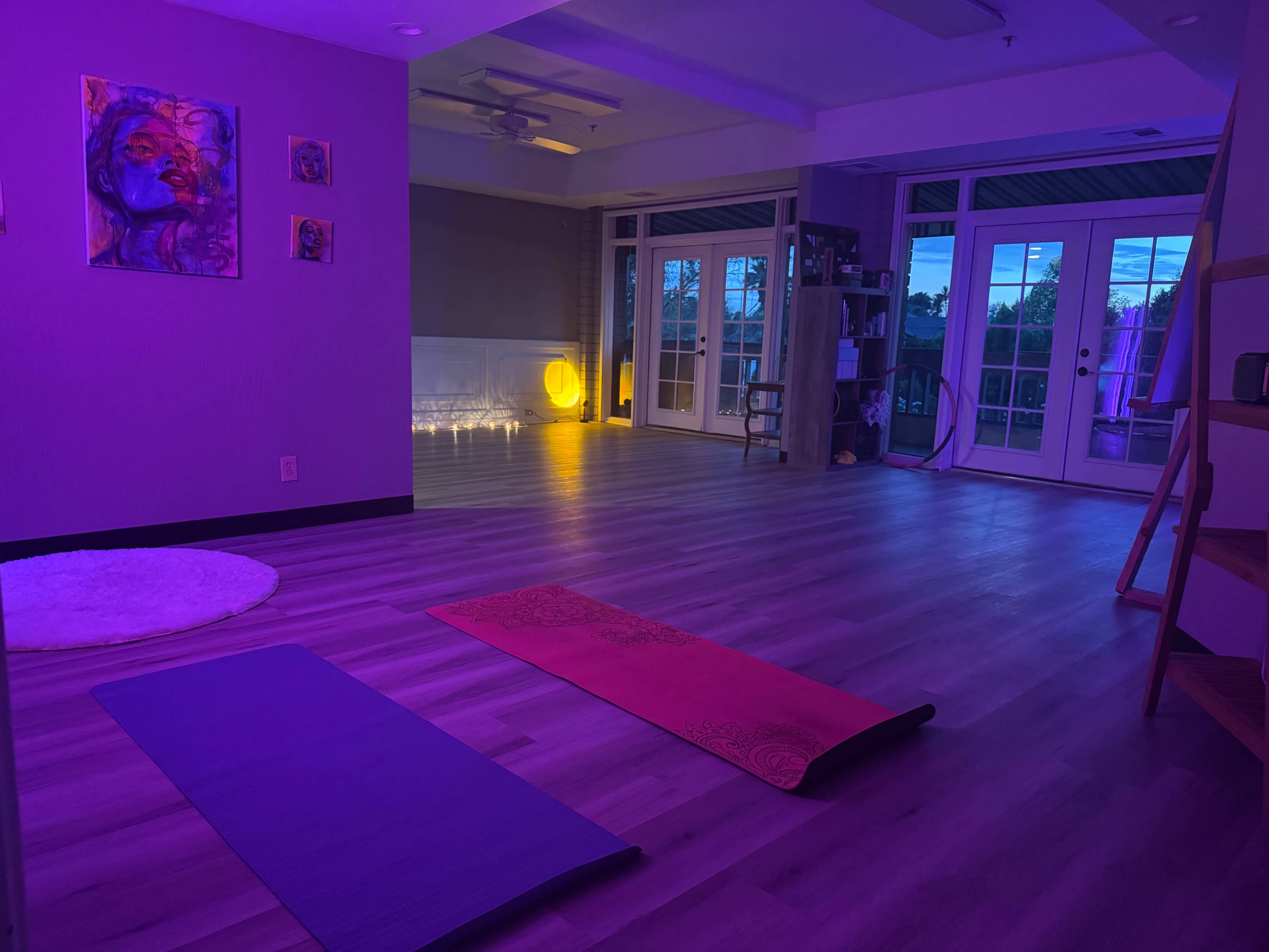 The image shows a dimly lit yoga studio featuring two exercise mats on the floor, surrounded by softly glowing lights and large windows.