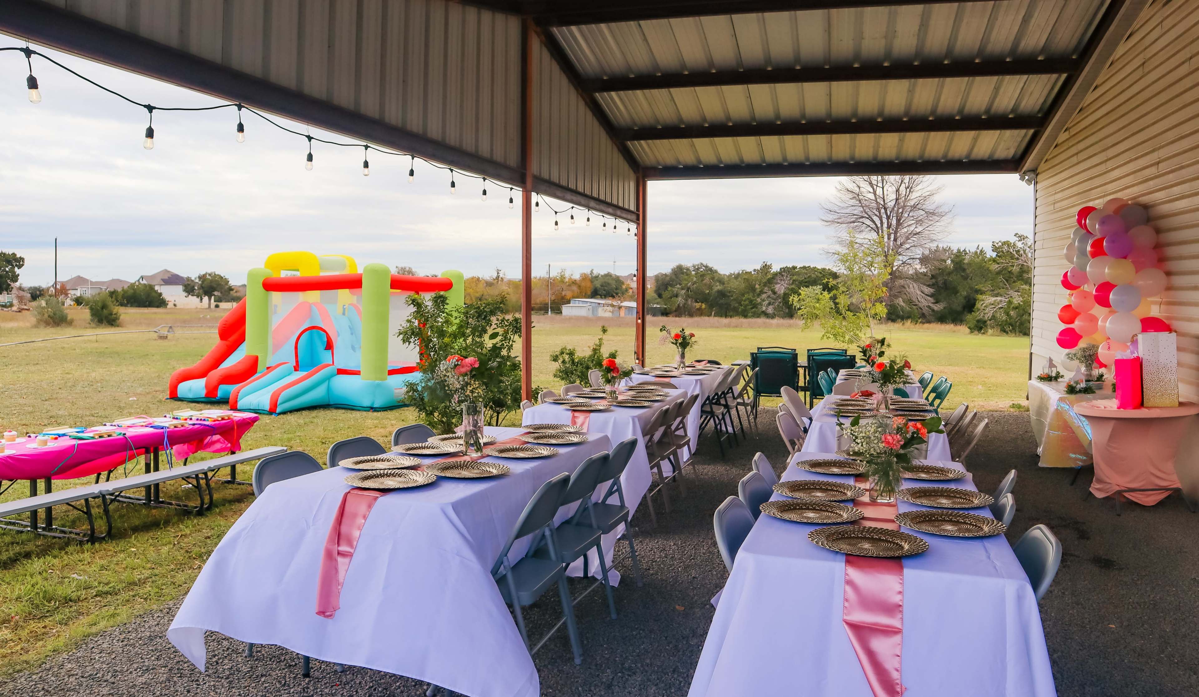 A covered outdoor area features rows of tables set with plates and decorations, alongside a colorful inflatable bounce house.