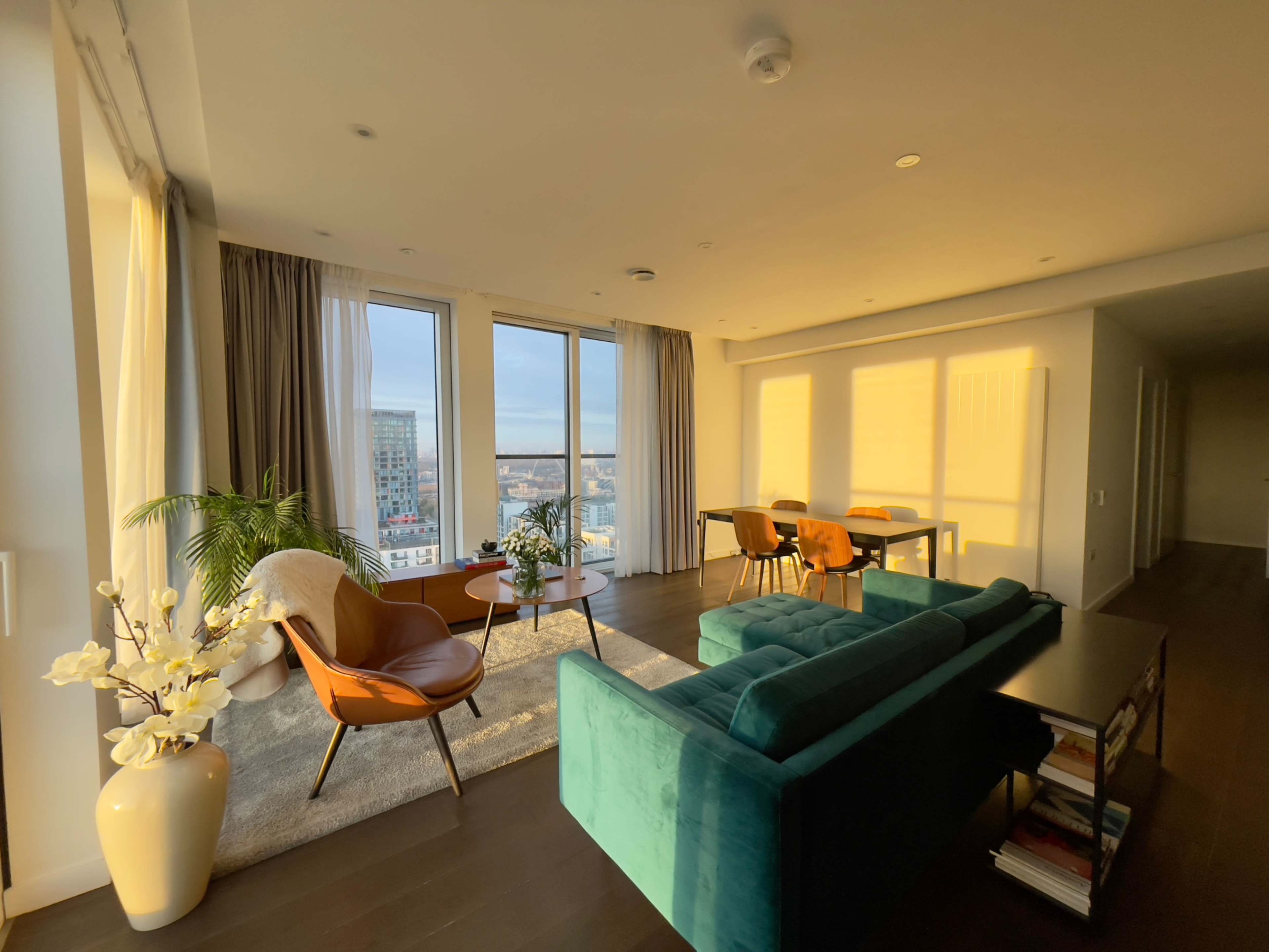 A modern living room features a teal sofa, brown armchairs, a dining table, and large windows allowing natural light to fill the space.