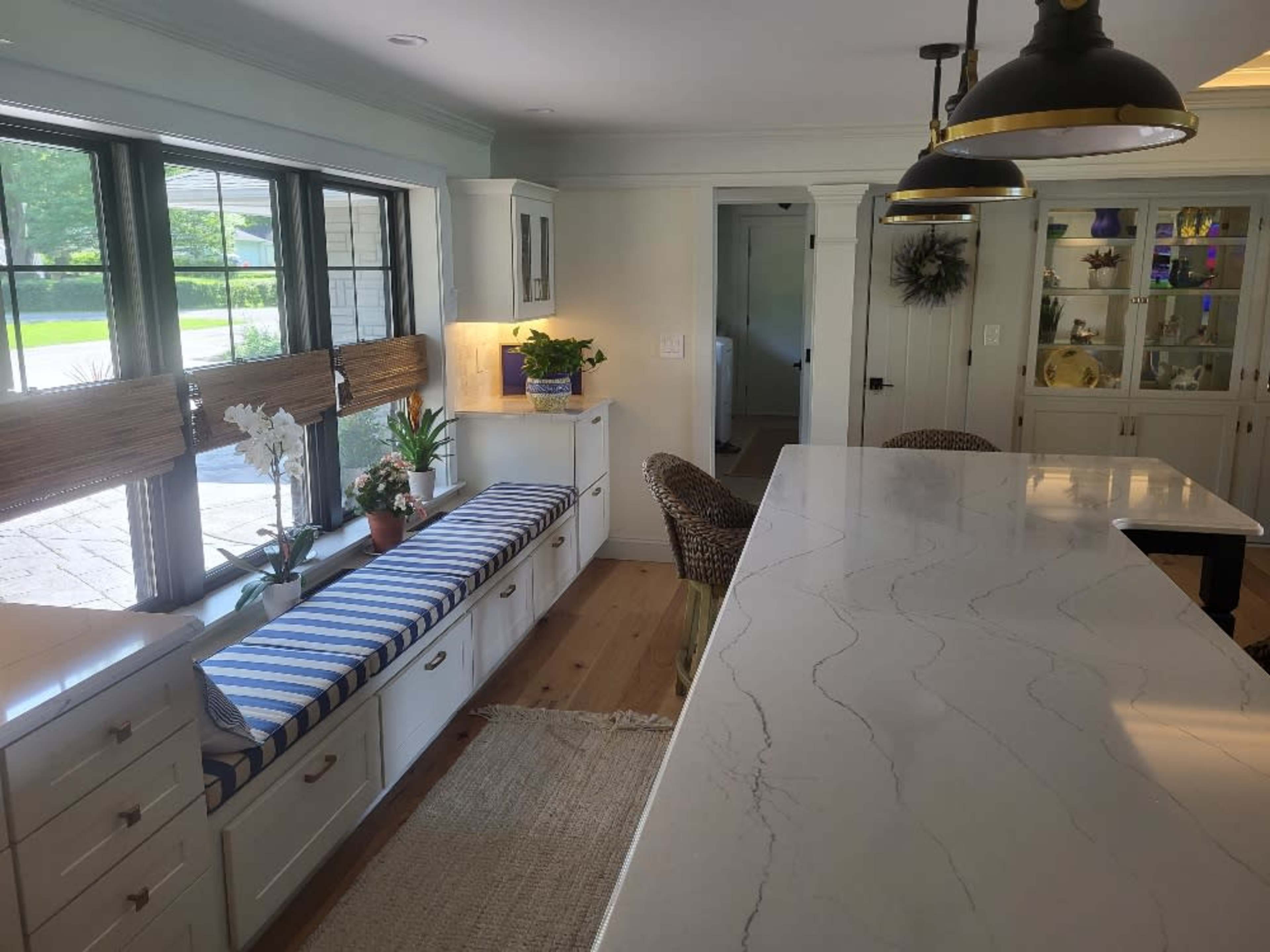 A modern kitchen with a large island, white cabinetry, a window seat with blue and white striped cushions, and pendant lighting above.