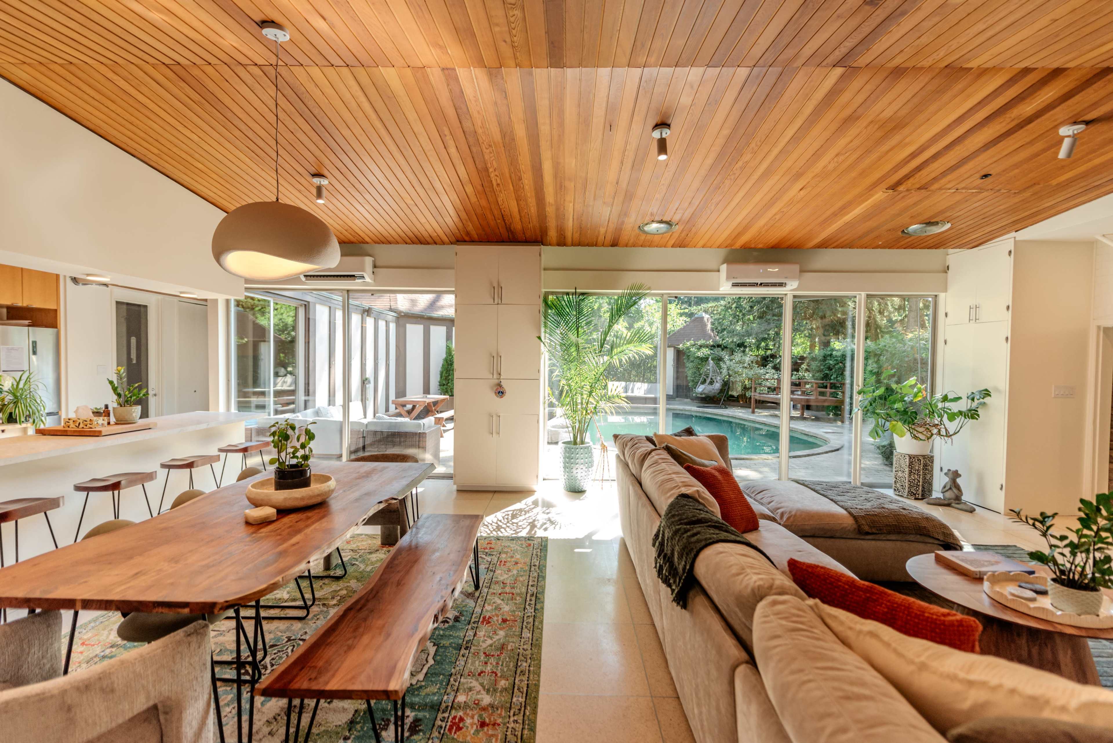 A spacious living area features a wooden ceiling, a large dining table, a sectional sofa, and glass doors leading to a pool area surrounded by greenery.