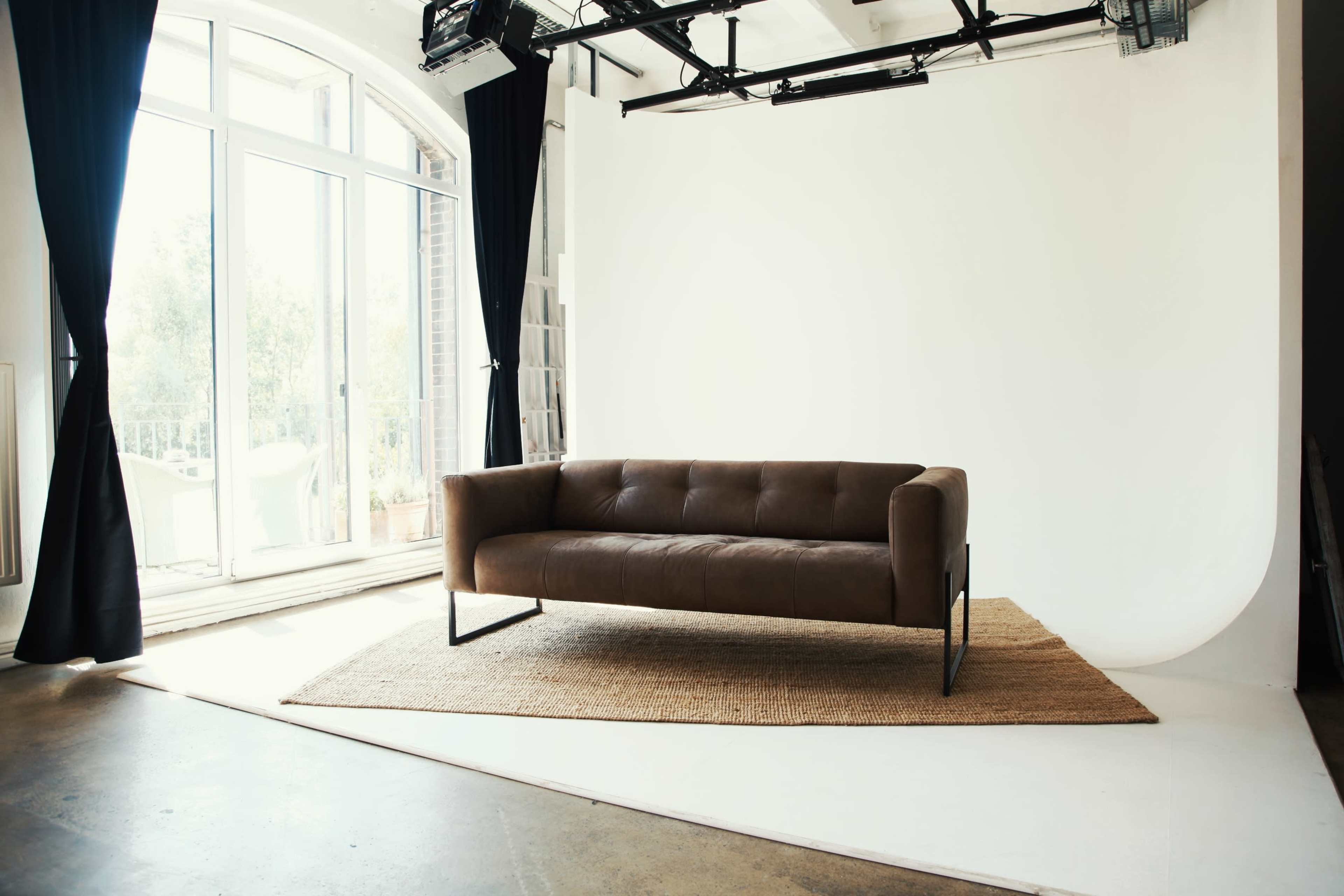 The image shows a modern studio space featuring a brown leather sofa placed on a jute rug in front of a white backdrop, with large windows letting in natural light.